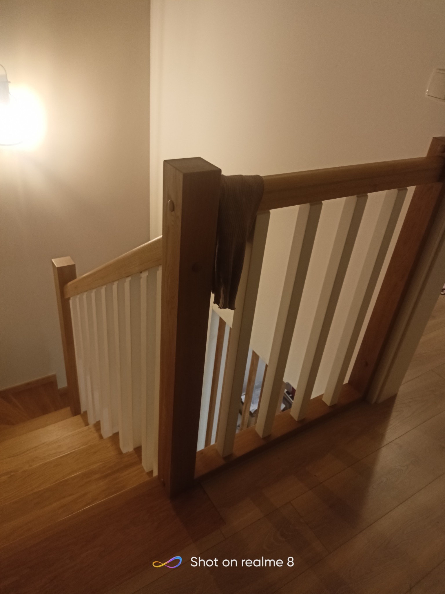 Widok z góry na drewniane schody z białymi balustradami, oświetlone ciepłym światłem, z elementem garderoby zawieszonym na poręczy.