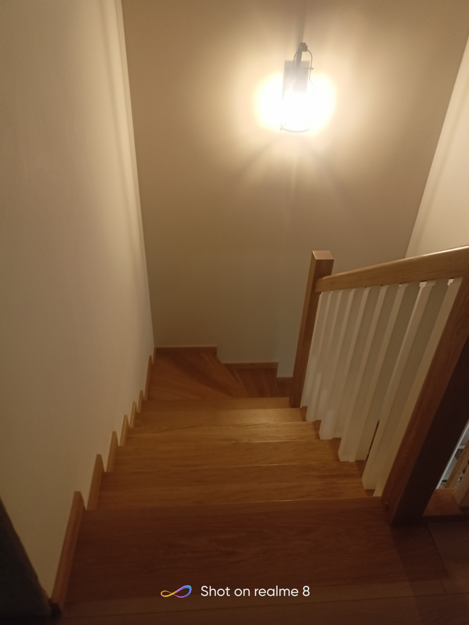 Widok z góry na drewniane schody z białymi balustradami, oświetlone przez lampę sufitową, ukazujące ciepłą kolorystykę drewna i prosty design.