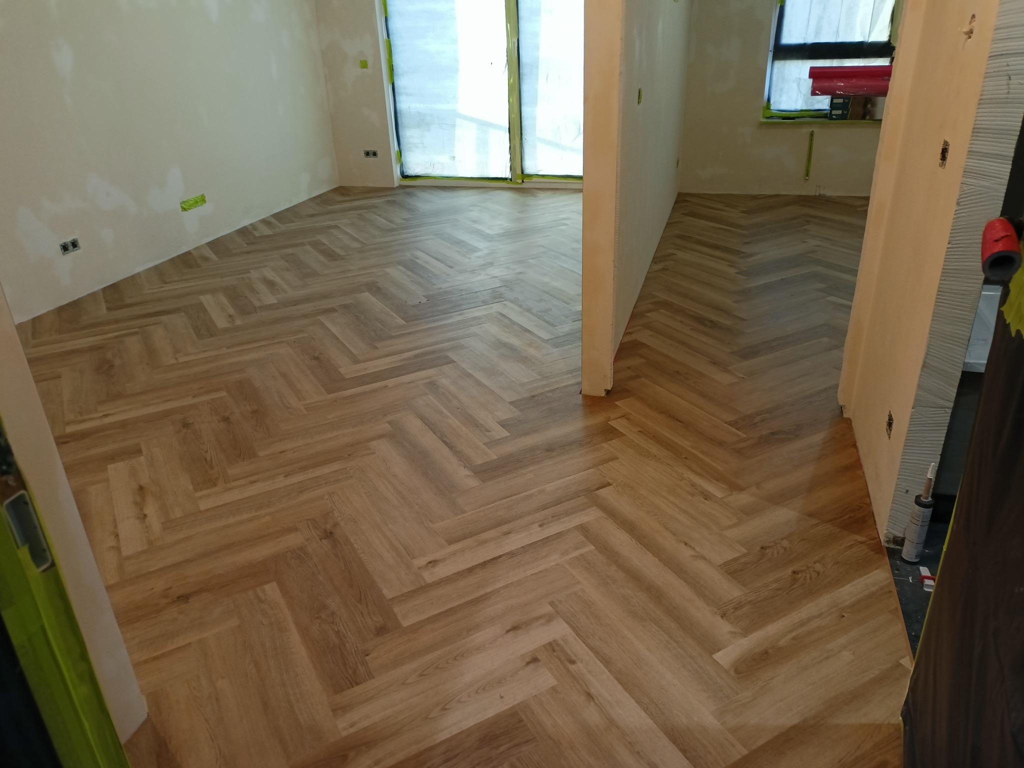 Ułożona podłoga z paneli w jodełkę w jasnym, pustym pokoju z oknem zabezpieczonym folią malarską, widoczne ślady po gładzi na ścianach.