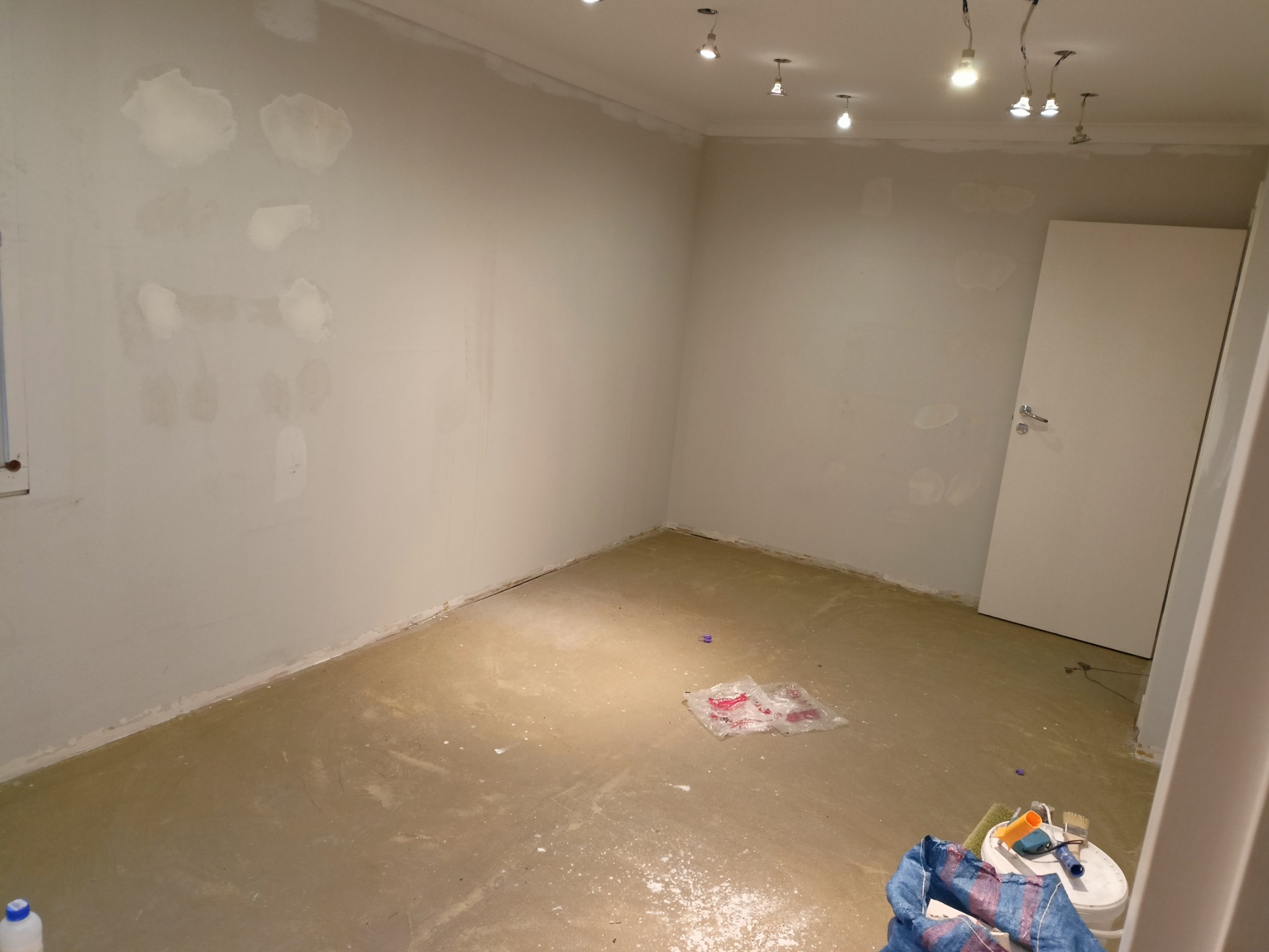 Pokój w trakcie przygotowania do malowania: ściany z widocznymi łatami po szpachlowaniu, surowa podłoga, widoczne narzędzia malarskie i zakurzone otoczenie.