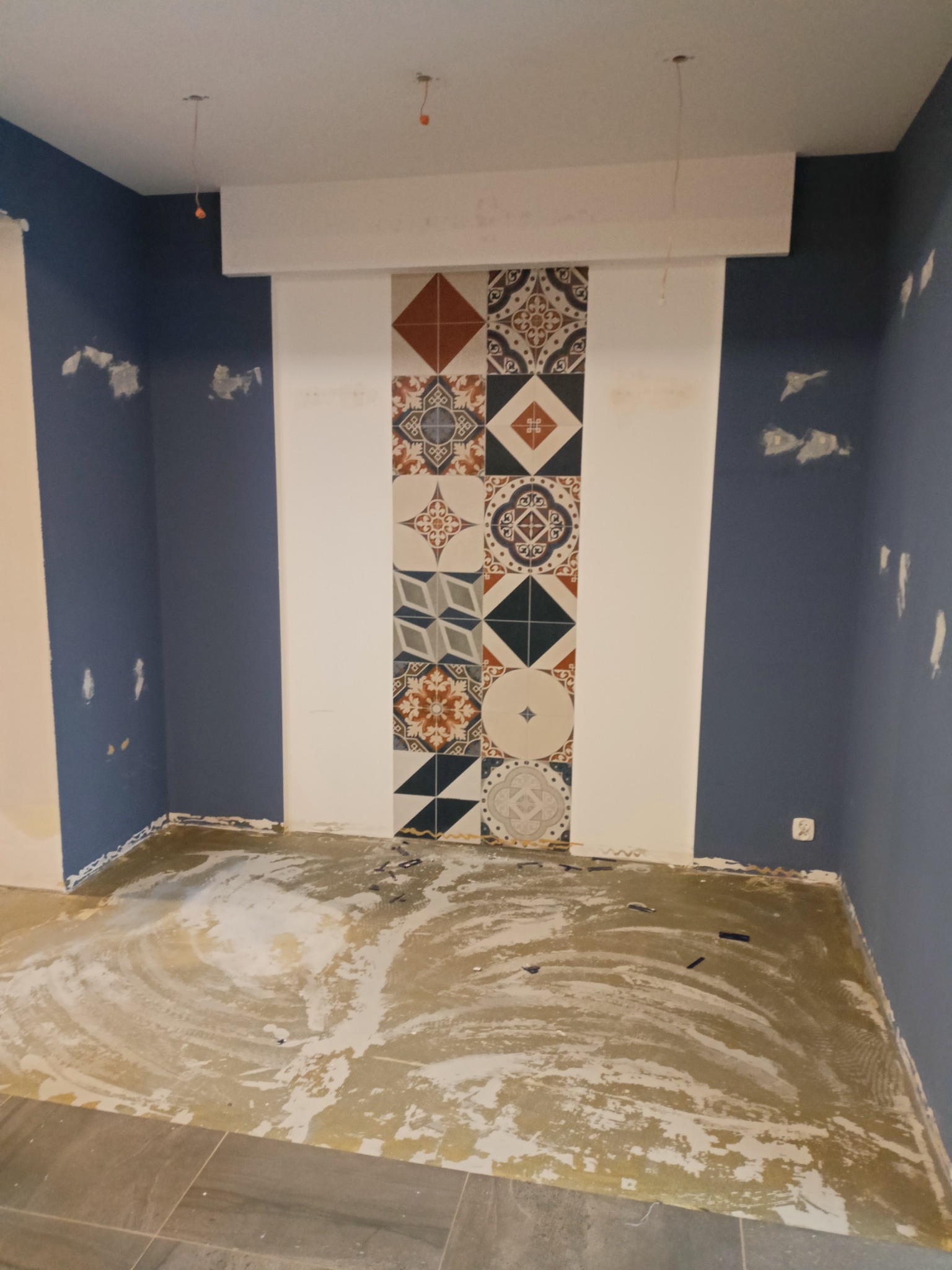 Wnętrze pokoju w trakcie remontu, z niebieskimi ścianami, fragmentem dekoracyjnej ściany z płytek w stylu vintage i przygotowanym podłożem pod nową podłogę.