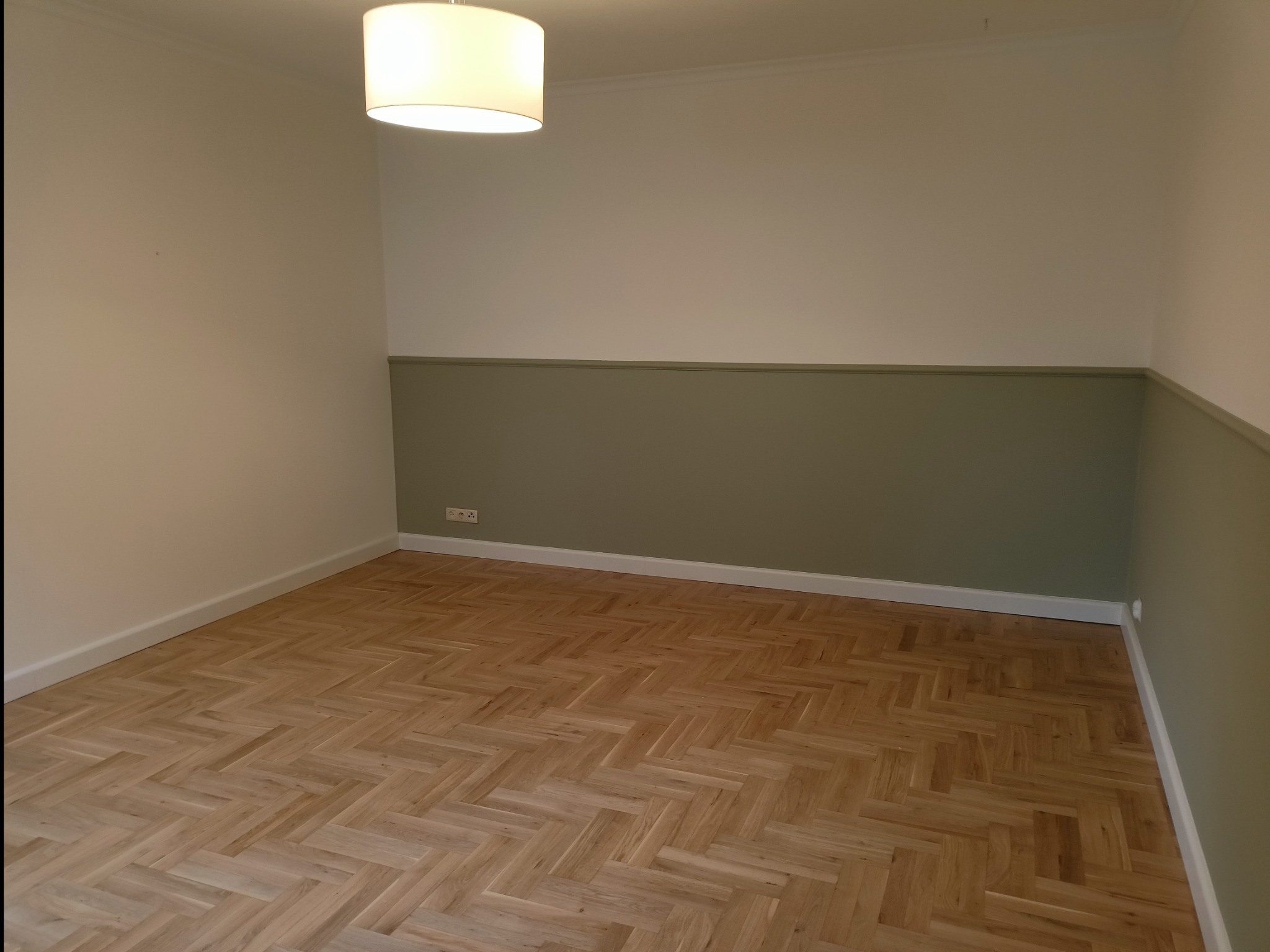 Pusty pokój po remoncie z parkietem ułożonym w jodełkę, ściany pomalowane na kremowo i oliwkowo, widoczne gniazdka elektryczne.