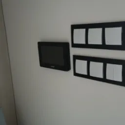 Panel sterowania alarmem Satel oraz dwa panele z białymi włącznikami światła w czarnych ramkach, zamontowane na białej ścianie.