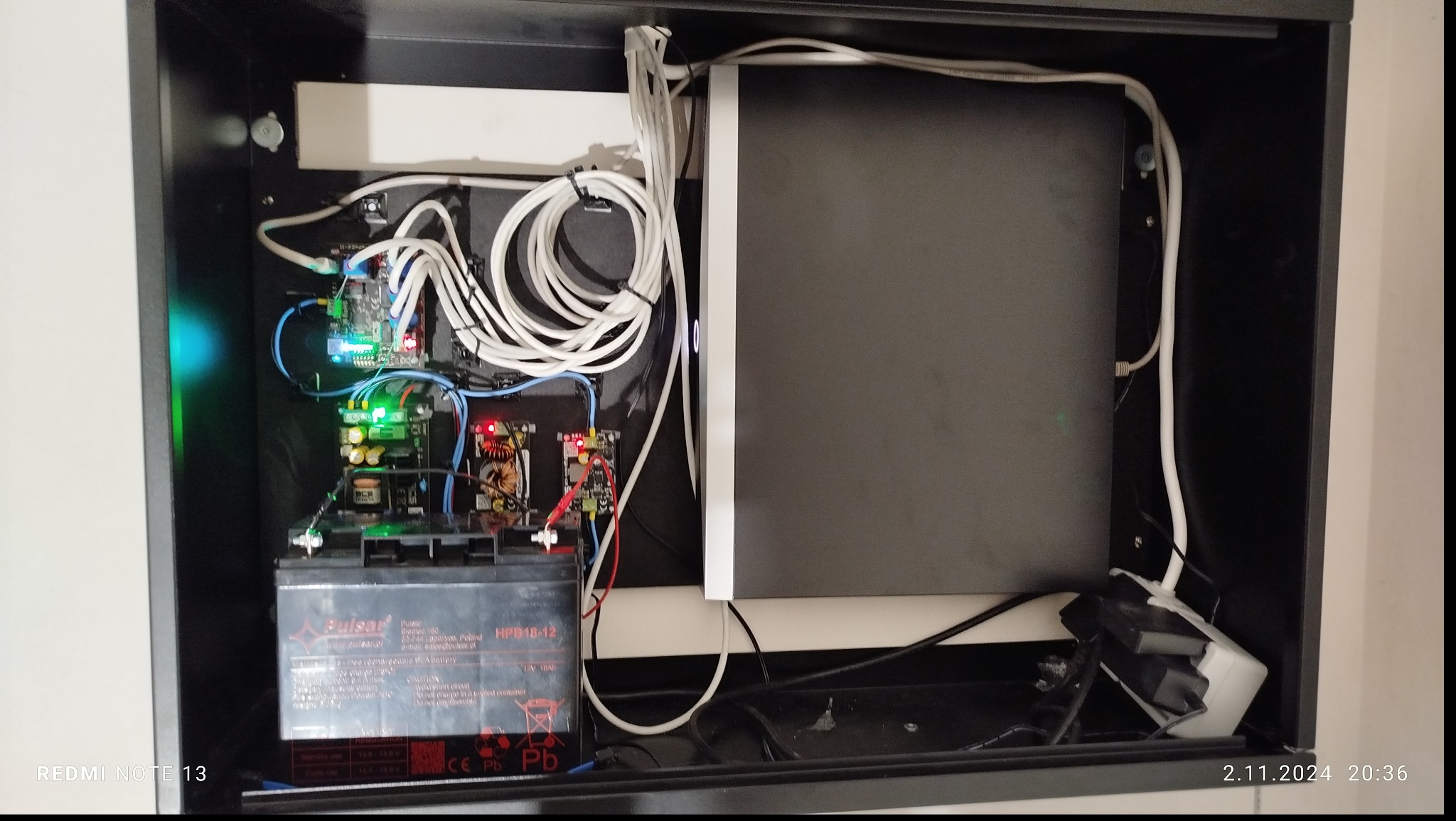 Wnętrze szafy sterowniczej z akumulatorem, płytkami drukowanymi i plątaniną białych kabli. Widoczne diody LED sygnalizujące pracę urządzeń.