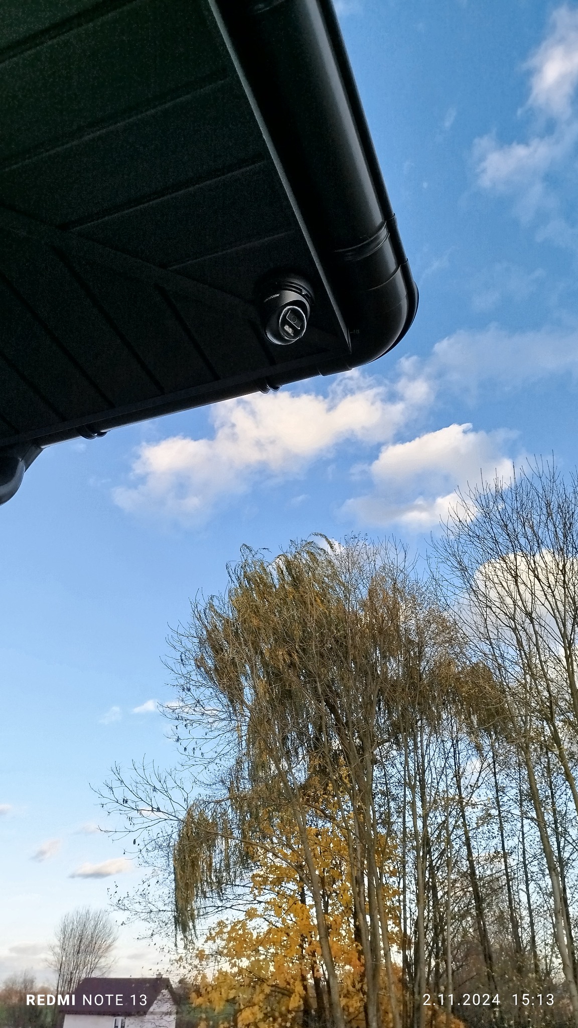 Kamera monitoringu kopułkowa zamontowana pod okapem dachu, widok z dołu na błękitne niebo z chmurami i drzewa jesienią.