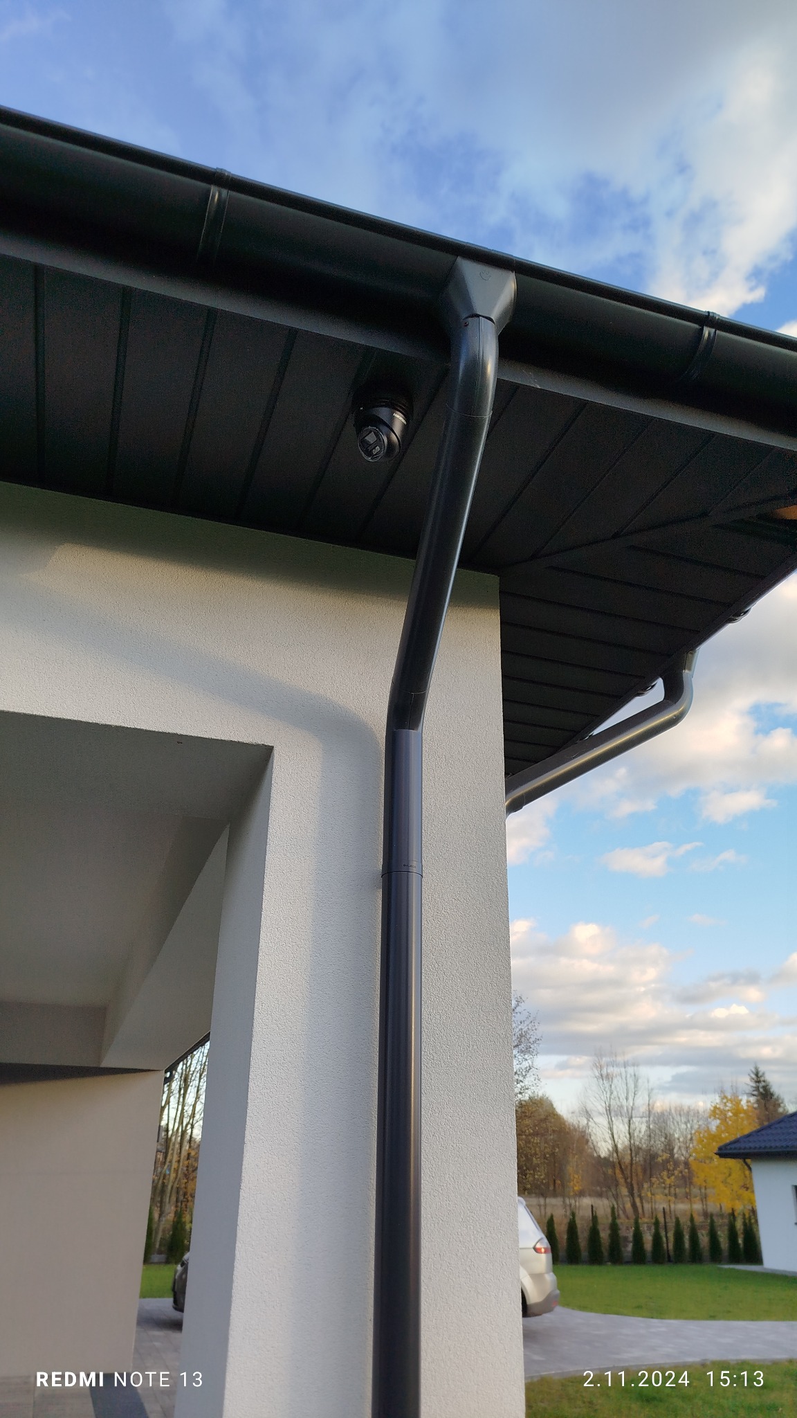 Zewnętrzna kamera monitoringu przymocowana do podbitki dachowej nowoczesnego domu z widoczną rynną spustową, ujęcie z dołu w słoneczny dzień.