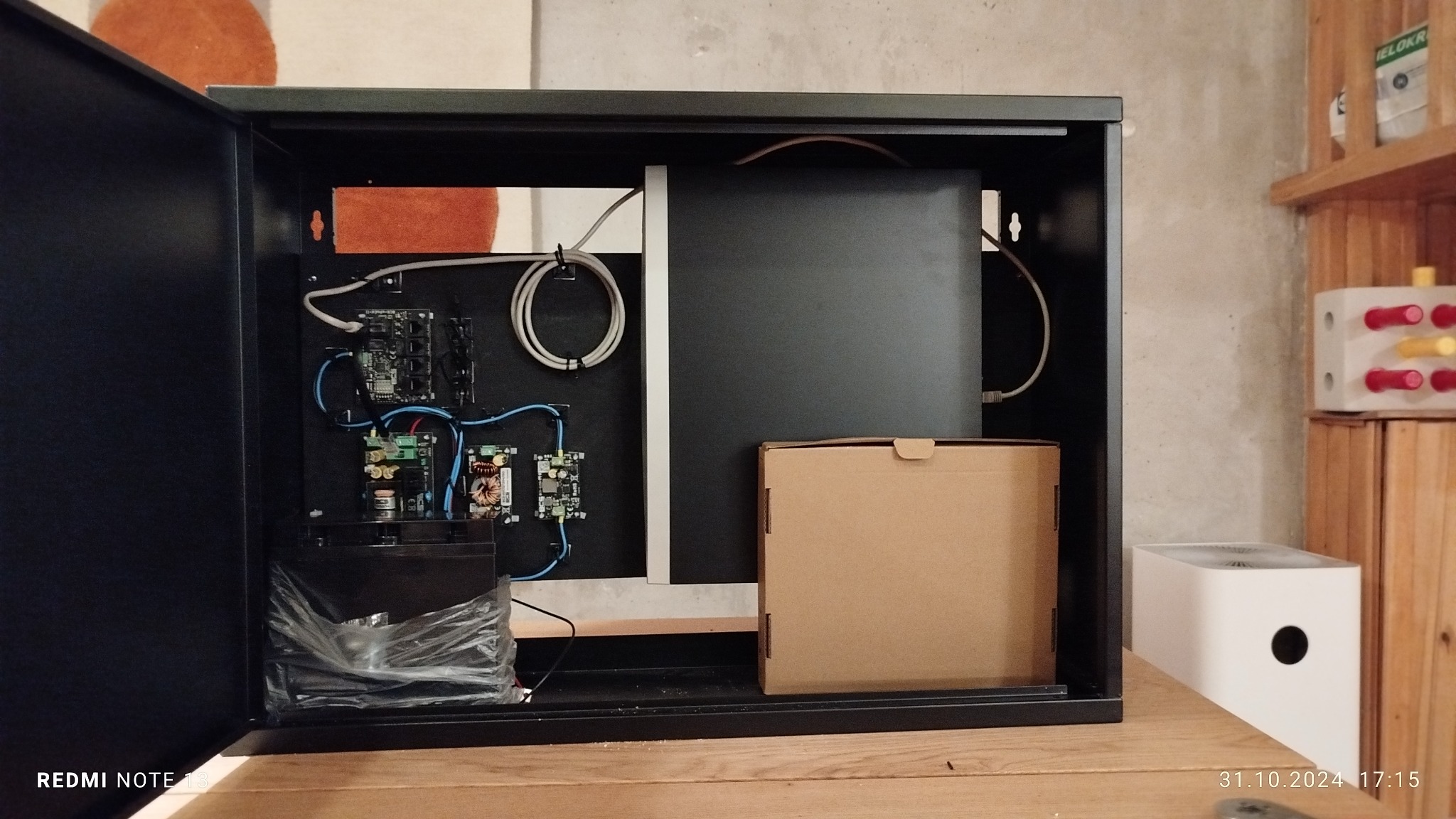 Otwarta czarna szafka z zamontowanymi modułami elektronicznymi, okablowaniem, akumulatorem w folii i kartonowym pudełkiem wewnątrz, na drewnianym blacie.