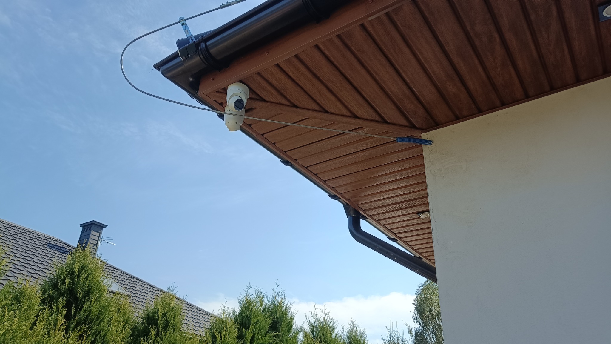 Kamera monitoringu zamontowana pod okapem budynku, widoczne przewody i elementy montażowe na tle błękitnego nieba.