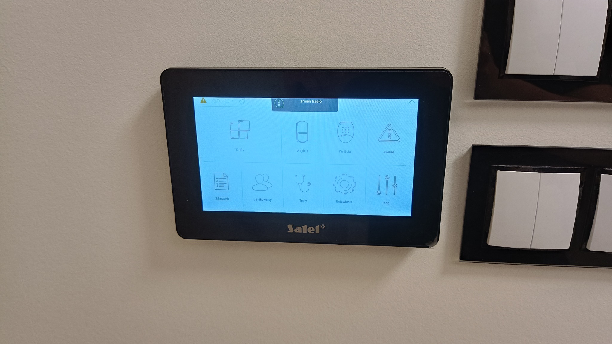 Ekran dotykowy systemu alarmowego Satel zamontowany na ścianie, obok widoczne włączniki światła w czarnej ramce. Na ekranie interfejs użytkownika z ikonami stref, wejść, wyjść, awarii, zdarzeń...