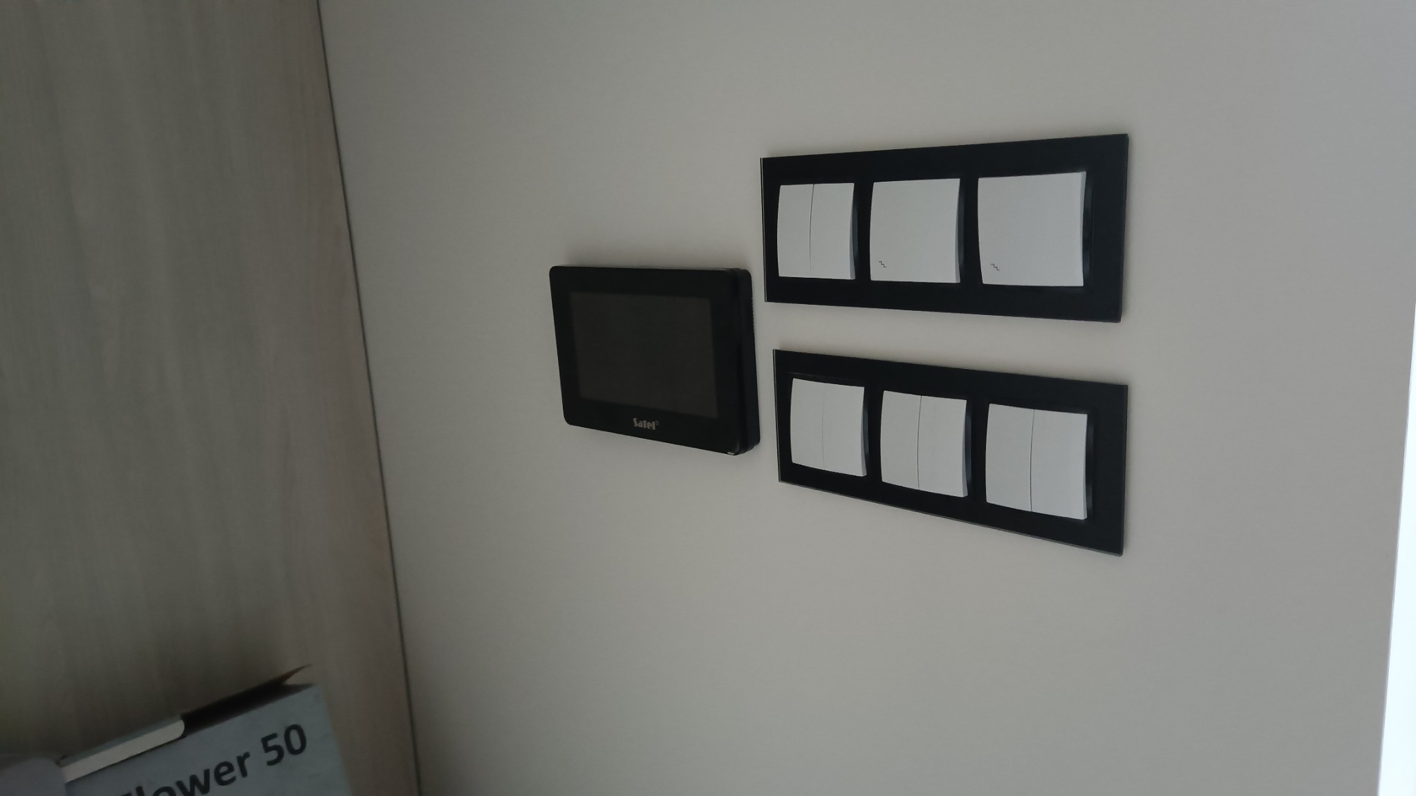 Panel sterowania alarmem Satel oraz dwa panele z białymi włącznikami światła w czarnych ramkach, zamontowane na białej ścianie.