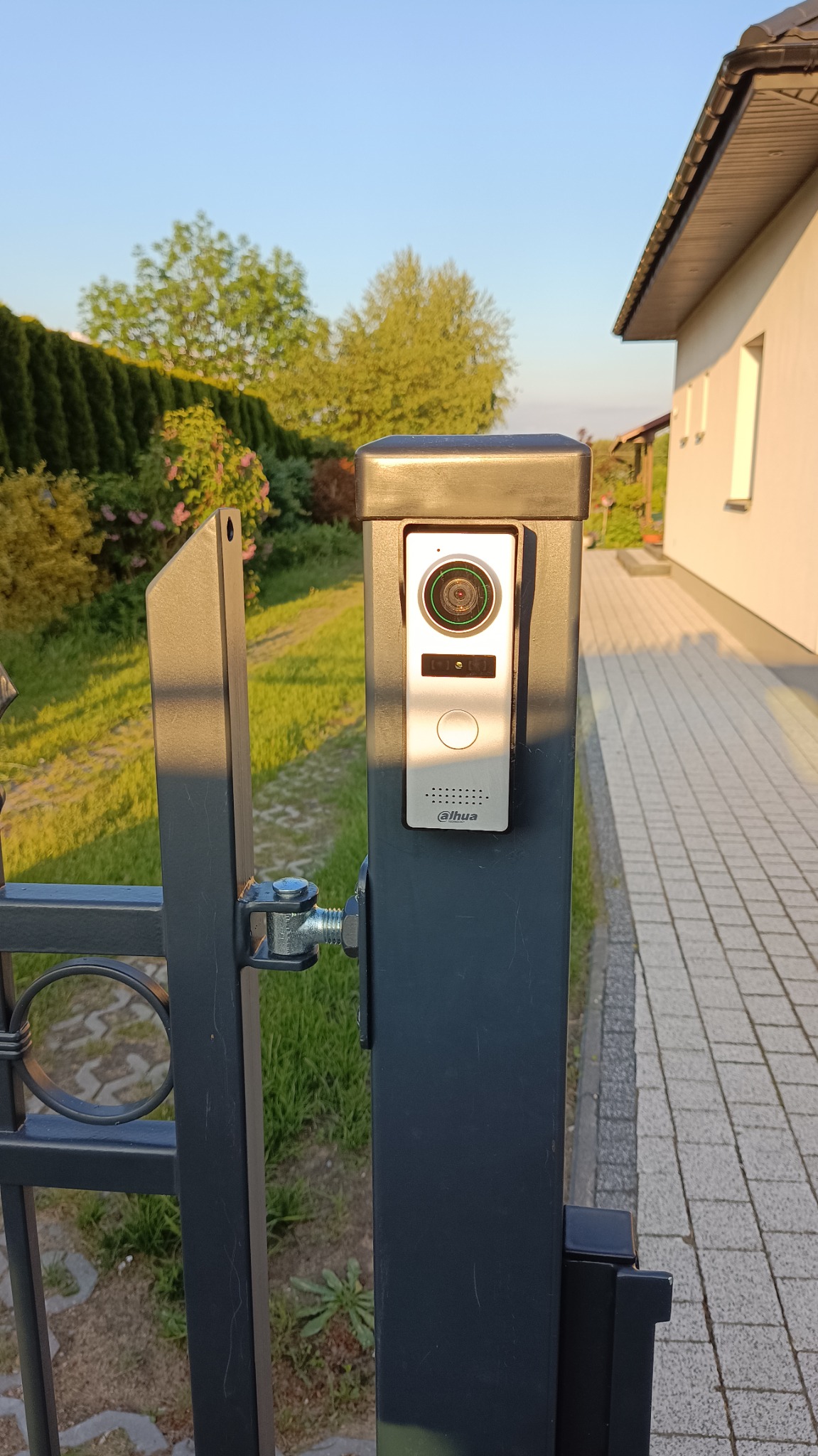 Domofon z kamerą marki Dahua zamontowany na metalowym słupku bramy wjazdowej, widoczne okablowanie, w tle trawnik i budynek mieszkalny w słoneczny dzień.