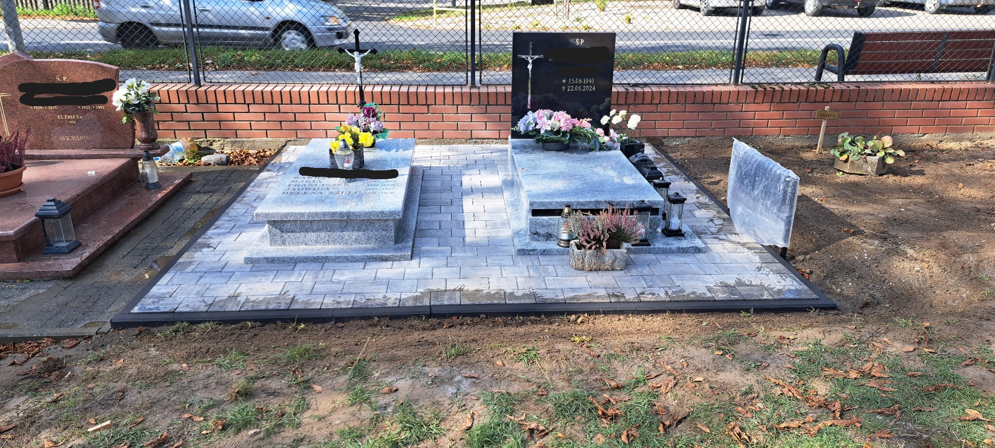 Wykończone miejsce pochówku z szarej kostki brukowej, z czarnymi obrzeżami i pomnikami. Widok na nagrobki, kwiaty i znicze. Tło stanowi ceglany mur i metalowe ogrodzenie.