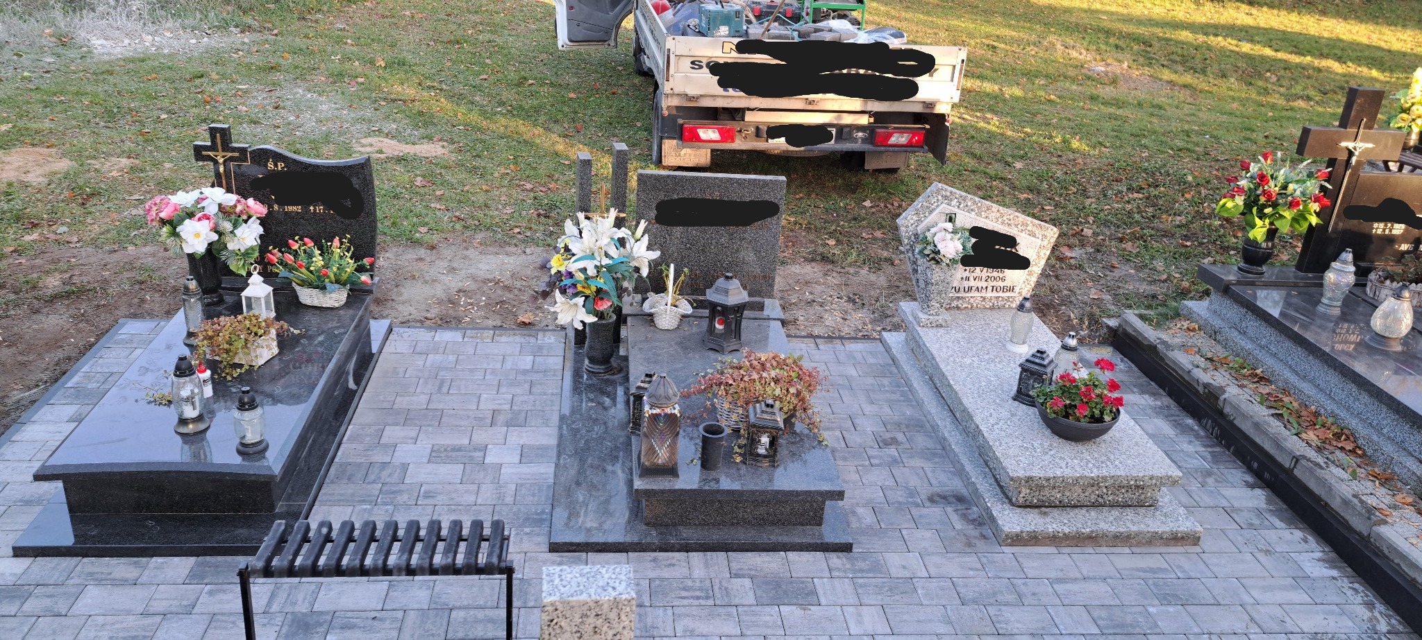 Świeżo ułożona, szara kostka brukowa na cmentarzu, otaczająca nagrobki z kwiatami i zniczami. W tle częściowo widoczny samochód dostawczy z narzędziami.