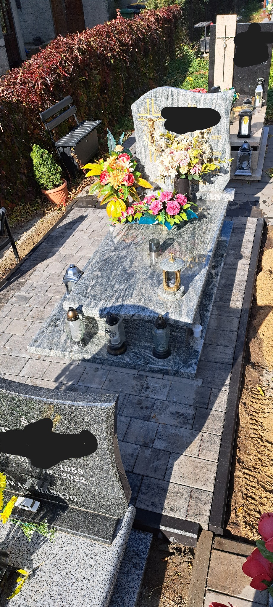 Granitowy nagrobek z kwiatami i zniczami, otoczony kostką brukową w odcieniach szarości. W tle widoczny fragment zieleni i ławka.