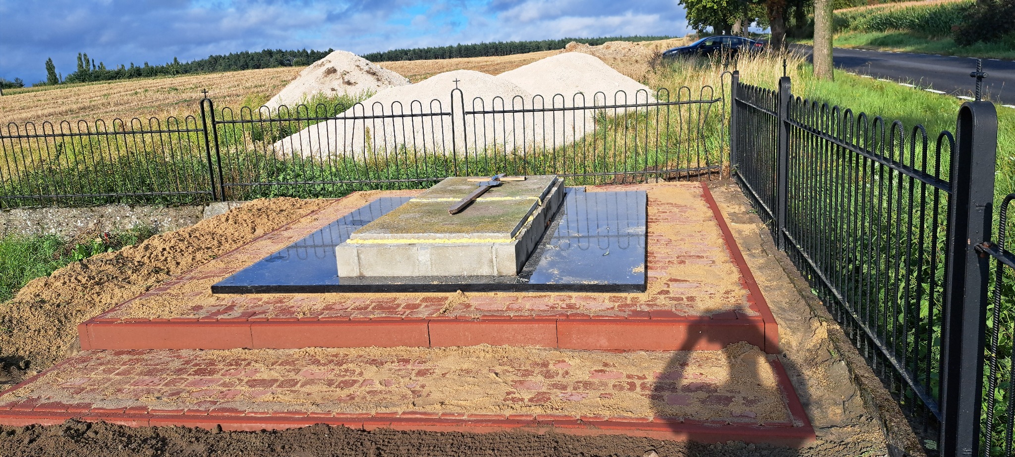 Wykończony nagrobek z czarnej płyty, otoczony czerwoną kostką i czarnym, metalowym ogrodzeniem. W tle widać stertę piasku i pola uprawne.