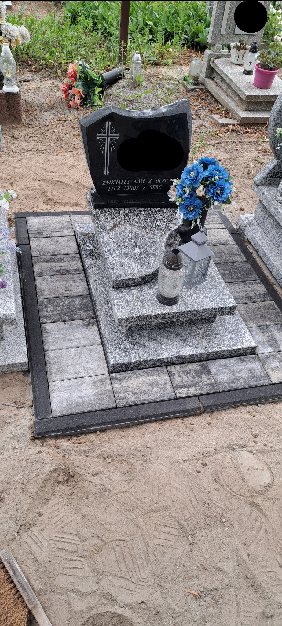 Granitowy nagrobek z czarną tablicą, otoczony szarymi kostkami brukowymi i czarnym obrzeżem, na piaszczystym podłożu. Kompozycja z kwiatów i lampion.