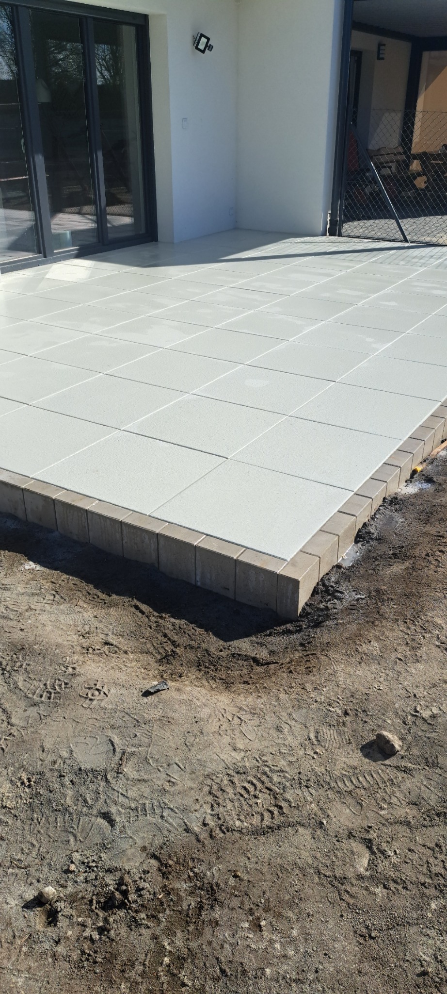 Wykończony taras z jasnych płyt betonowych obrzeżony kostką w kolorze beżowym, przylegający do białego budynku z dużymi oknami przesuwnymi.