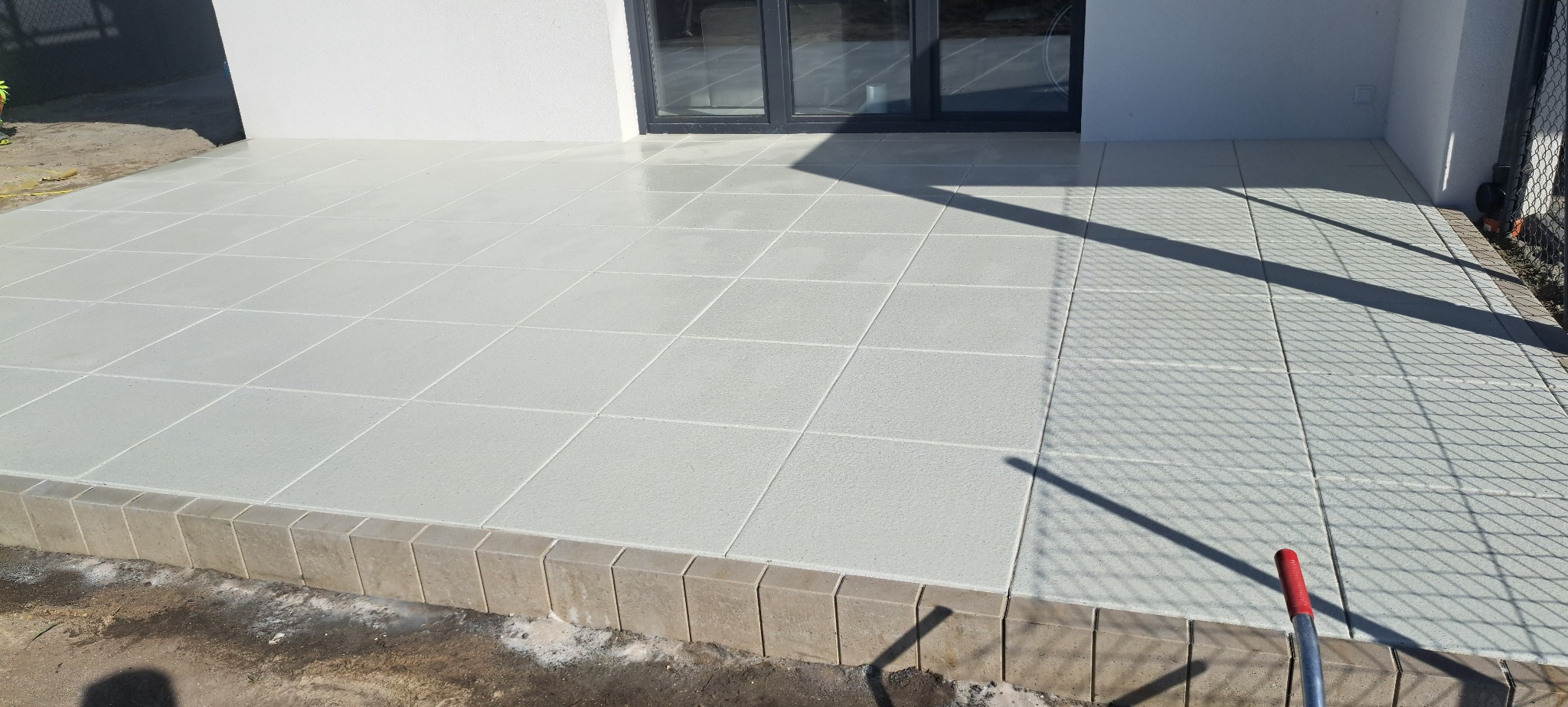 Wykończony taras z jasnych, kwadratowych płyt, z widocznymi fugami i obrzeżem z kostki. Cień rzucany przez ogrodzenie panelowe.