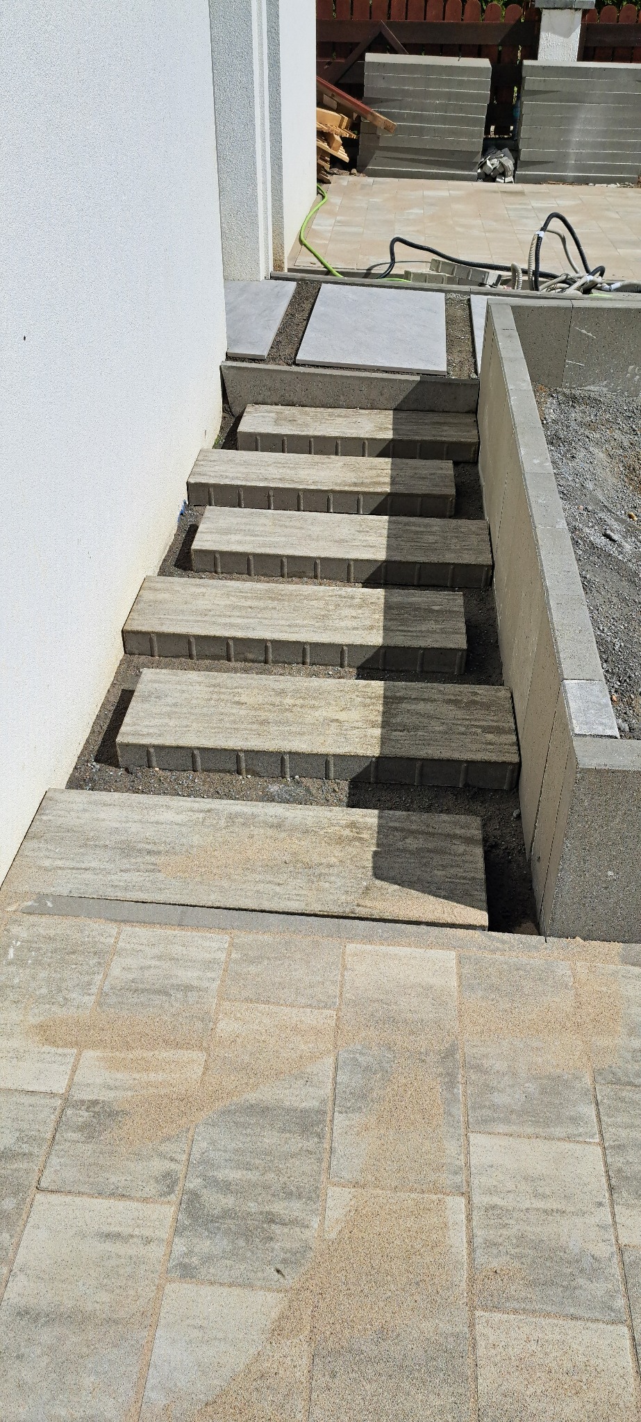 Schody z kostki brukowej prowadzące do podwyższonego tarasu, widok z góry, z jasną kostką na dole i zapasem materiału budowlanego w tle.