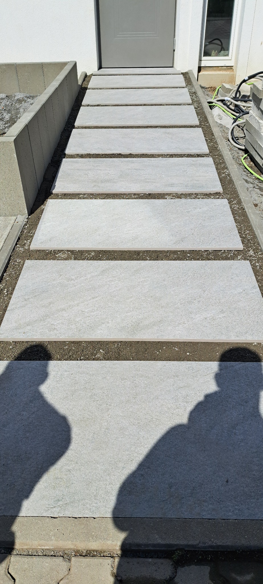 Nowoczesne, prostokątne płyty chodnikowe prowadzą do drzwi. Minimalistyczny design z widocznymi elementami przygotowania podłoża i cieniem osoby fotografującej.