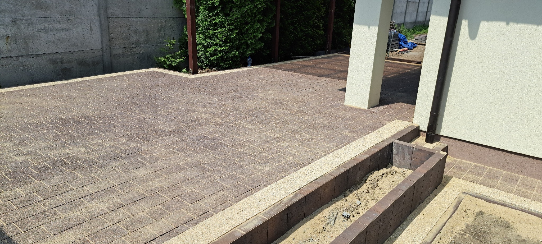 Ukończona nawierzchnia z kostki brukowej w Dąbrowie, wzór jodełki, obrzeża z jasnego żwiru. Widoczny fragment elewacji budynku i betonowe ogrodzenie.