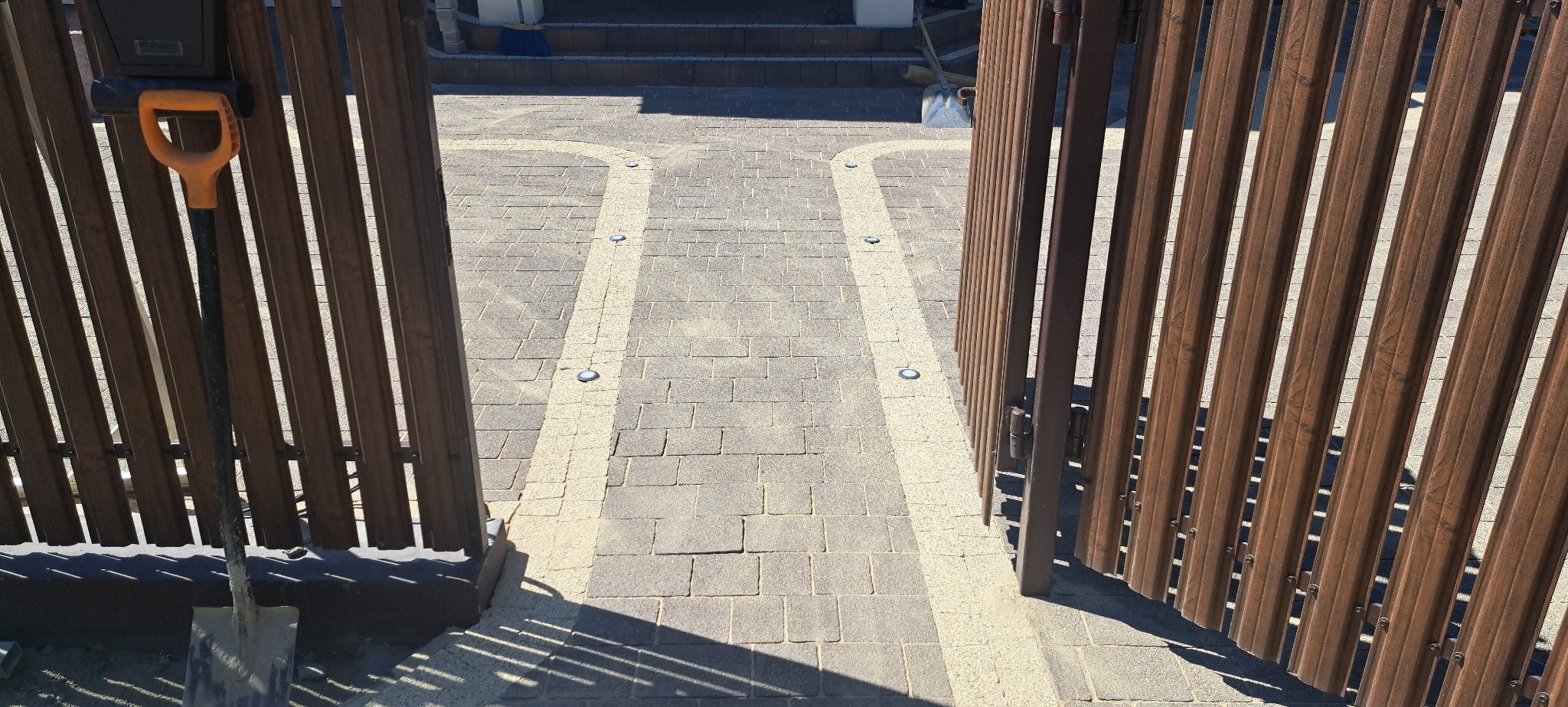 Otwarta, brązowa furtka panelowa, prowadząca na ścieżkę z kostki brukowej z oświetleniem punktowym. Z lewej strony oparta o ogrodzenie łopata.