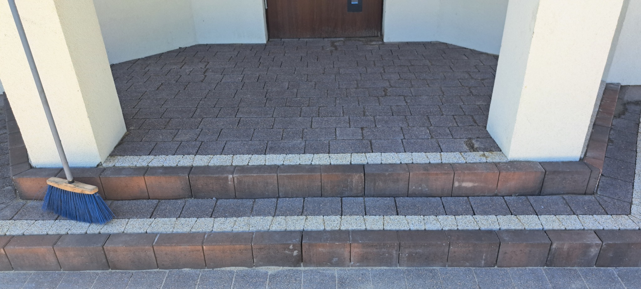Schody wejściowe wyłożone szarą kostką brukową z brązowym obramowaniem i elementami z jasnej kostki. Przy kolumnie oparta niebieska zmiotka.