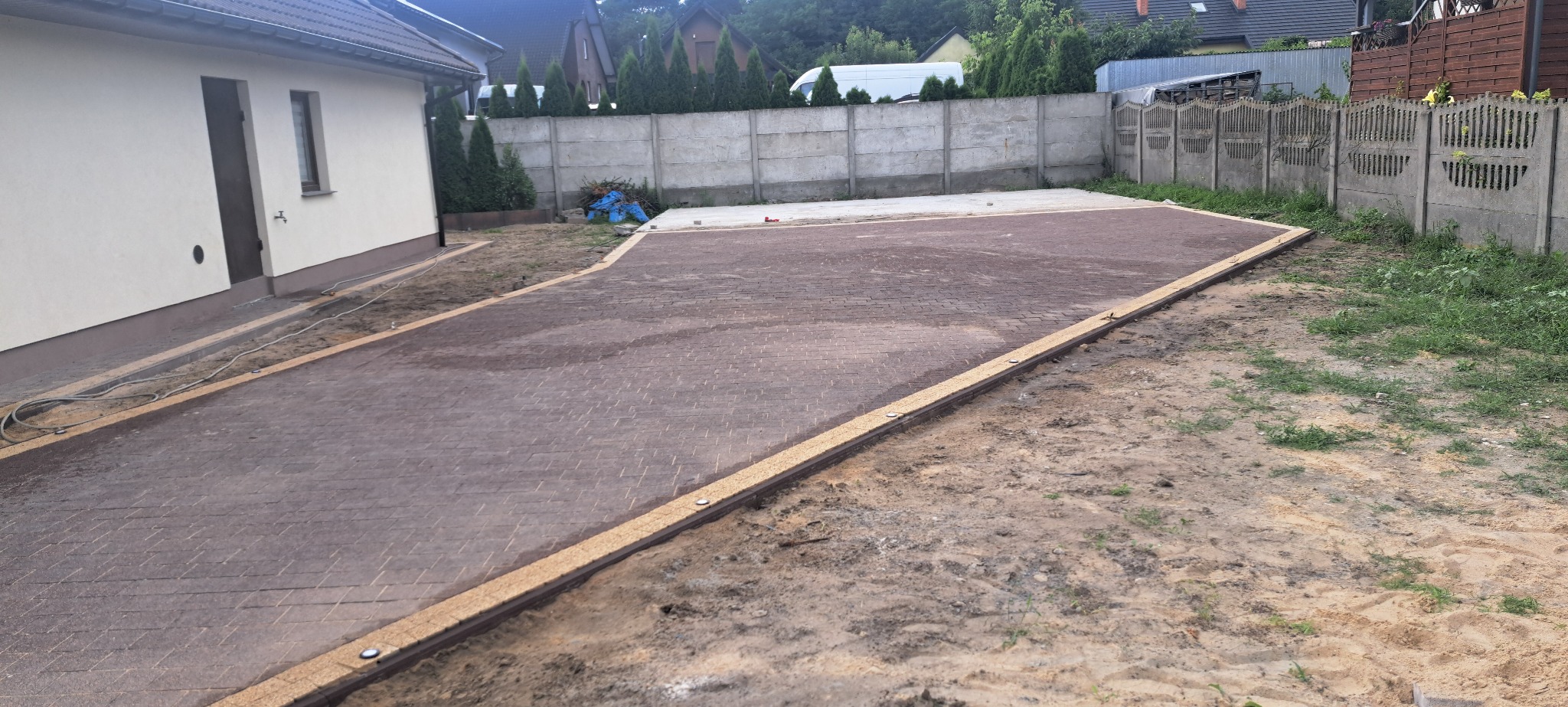 Ukończony podjazd z brązowej kostki brukowej ułożonej we wzór jodełki, z widocznymi obrzeżami i elementami oświetlenia, przylegający do budynku.