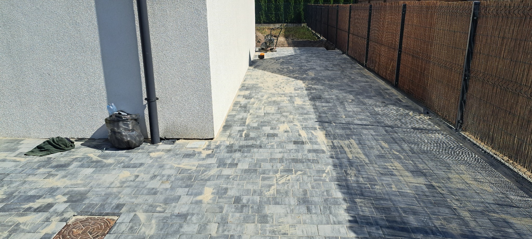 Ułożona szara kostka brukowa przy brązowym ogrodzeniu panelowym i ścianie budynku. Widoczny czarny worek na śmieci i właz kanalizacyjny. Częściowe zacienienie na kostce.