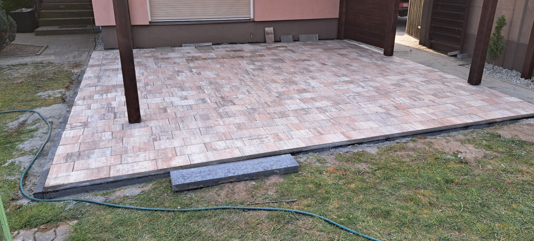 Ułożona, beżowo-brązowa kostka brukowa na podjeździe, widoczne obrzeża i słupy konstrukcji zadaszenia. Częściowo wykończona nawierzchnia.
