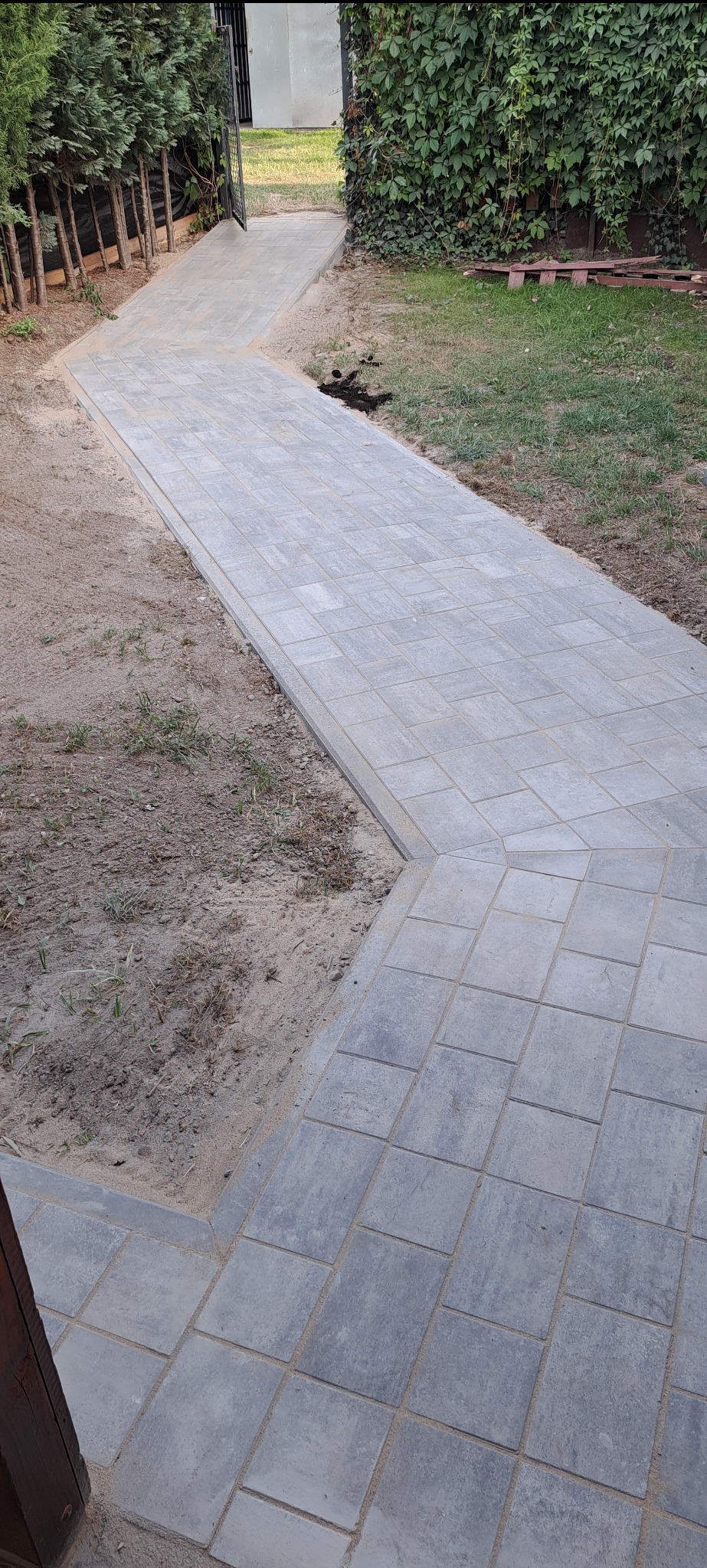 Ścieżka z szarej kostki brukowej prowadząca przez ogród, z widocznym trawnikiem i zielenią w tle. Kostka ułożona w regularny wzór.