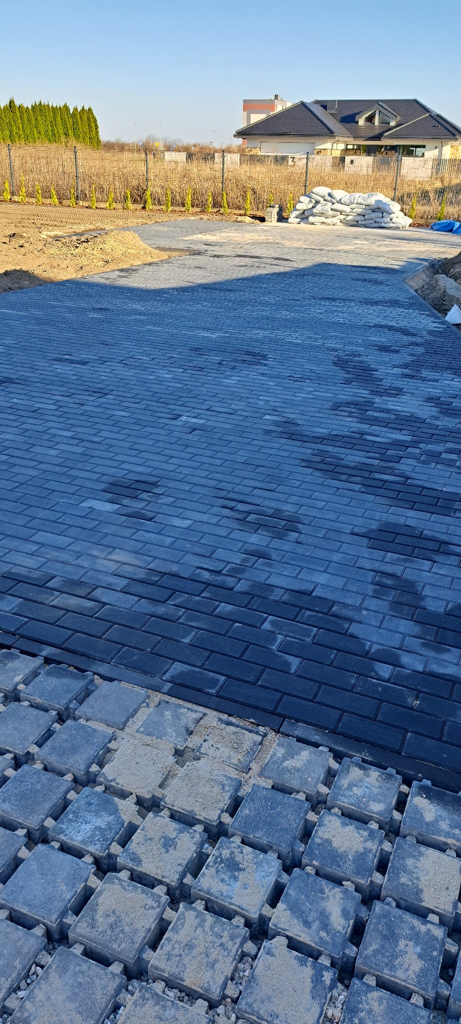 Ciemnoszara kostka brukowa ułożona na podjeździe, widoczne elementy podbudowy. W tle dom jednorodzinny i ogrodzenie z tujami.