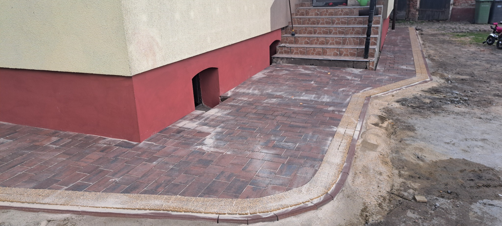 Ułożona kostka brukowa w odcieniach brązu przed budynkiem z wejściem do piwnicy. Obrzeża wykończone drobnym kamieniem. Widoczne schody wejściowe.