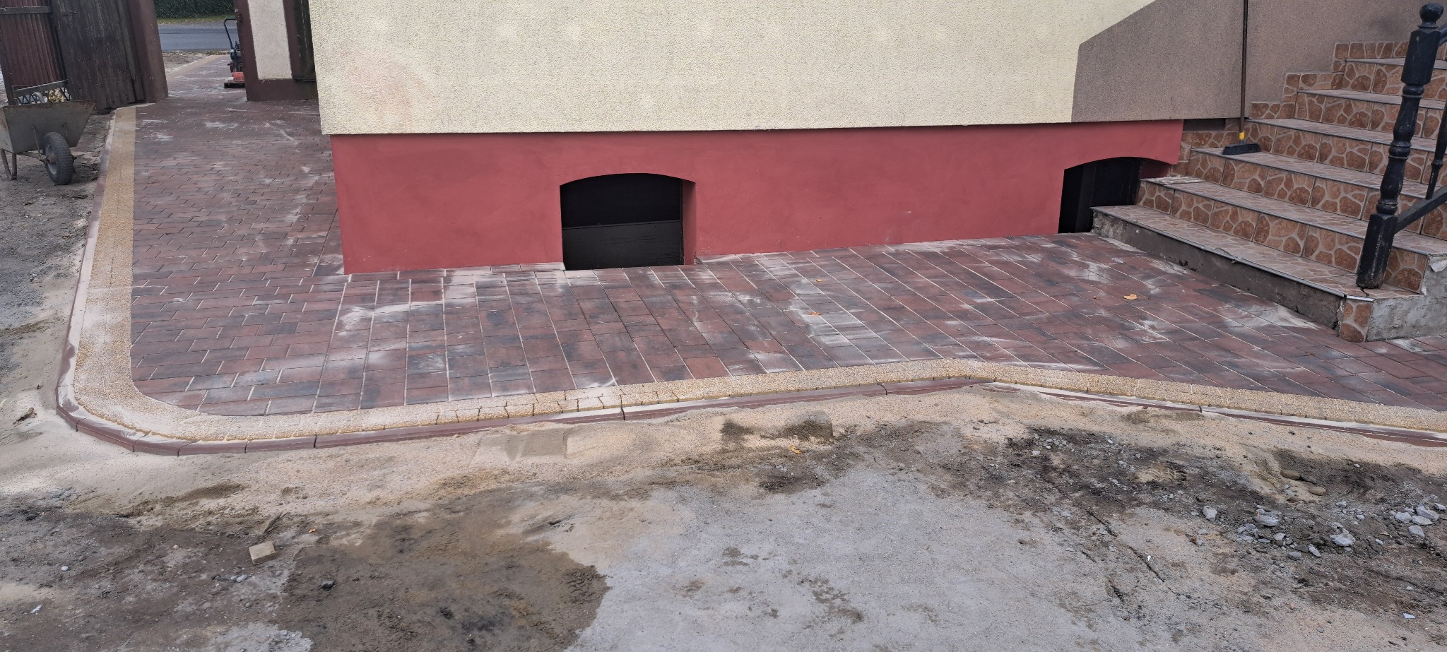 Ułożona kostka brukowa wzdłuż budynku z widocznymi schodami i elementami wykończeniowymi. Częściowo widoczne przygotowane podłoże.