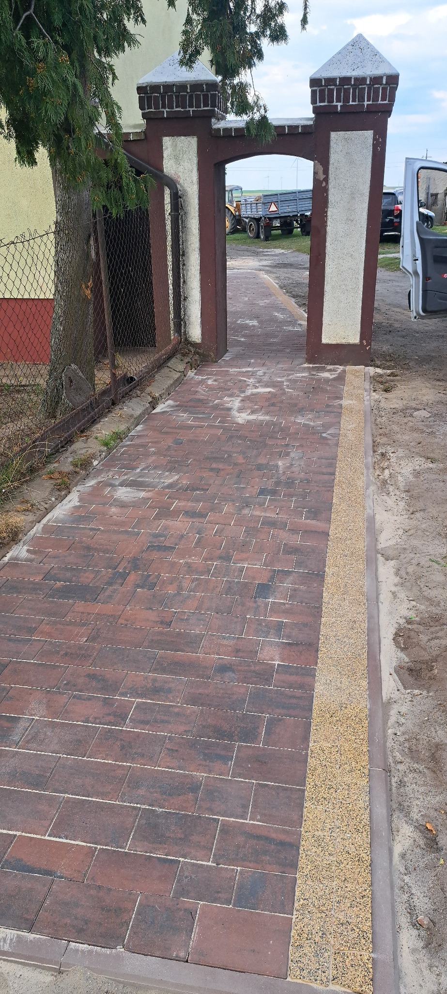 Ułożona, czerwono-brązowa kostka brukowa prowadząca przez bramę z ceglanymi słupkami. Krawężnik z jasnego kruszywa. W tle traktor z przyczepą i samochód.