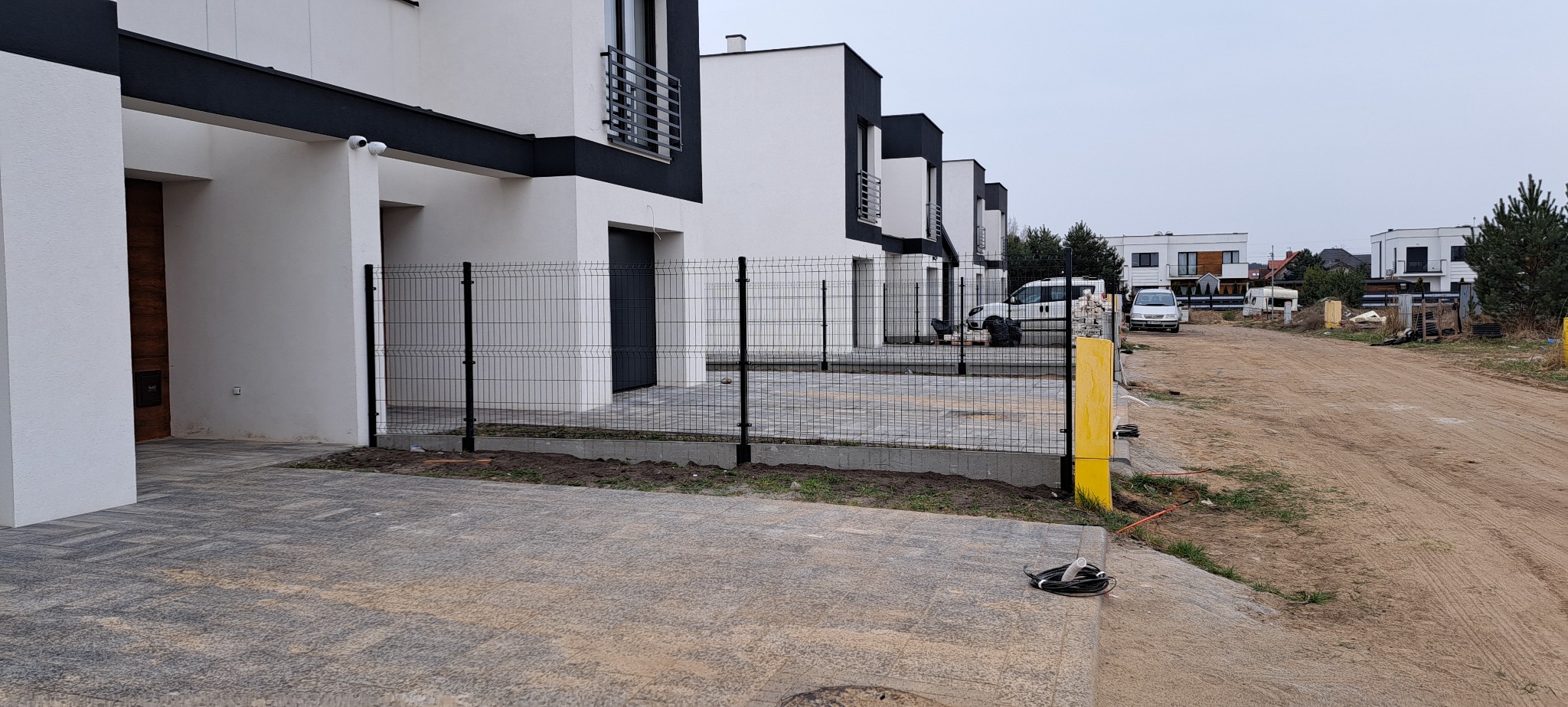 Nowoczesne ogrodzenie panelowe posesji w Dąbrowie. Czarne panele kontrastują z białą elewacją budynków. Widoczny fragment kostki brukowej i droga gruntowa.