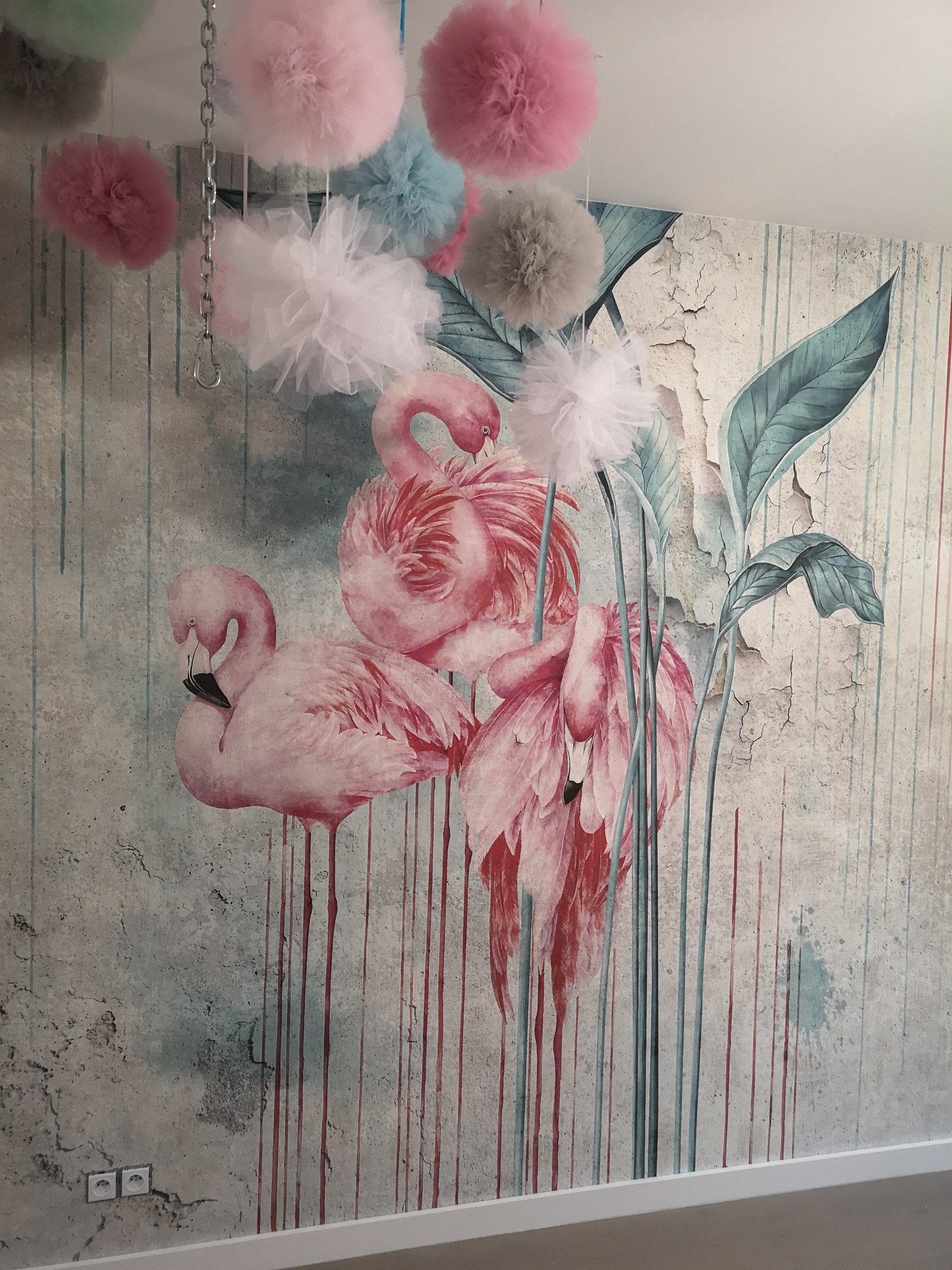 Tapeta z motywem różowych flamingów i tropikalnych liści na tle imitującym popękany mur, uzupełniona dekoracjami z tiulu w pastelowych kolorach zawieszonymi na łańcuchu.