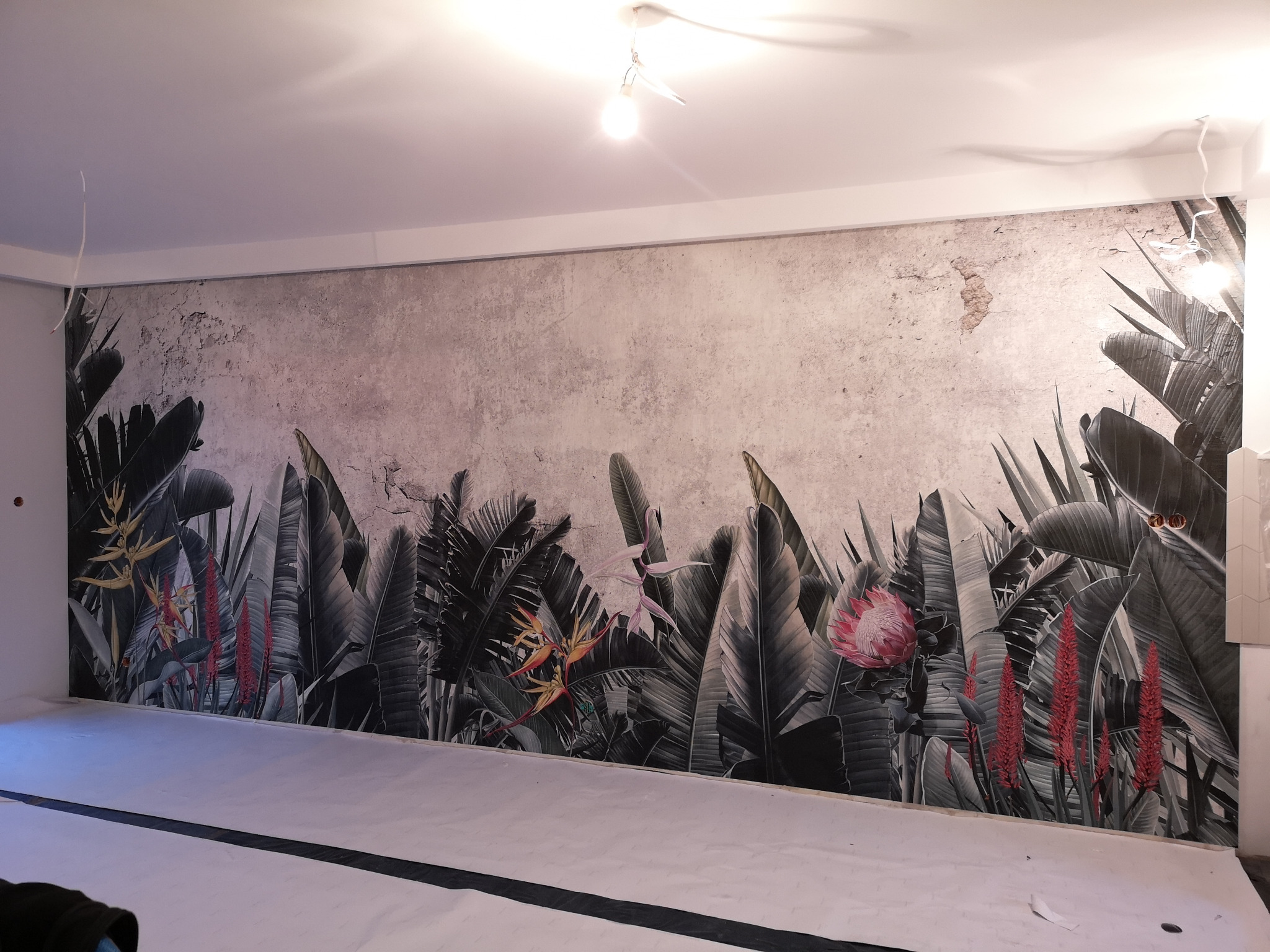Ściana po nałożeniu fototapety z motywem tropikalnych liści i kwiatów na tle imitującym betonową ścianę. Podłoga zabezpieczona folią malarską.