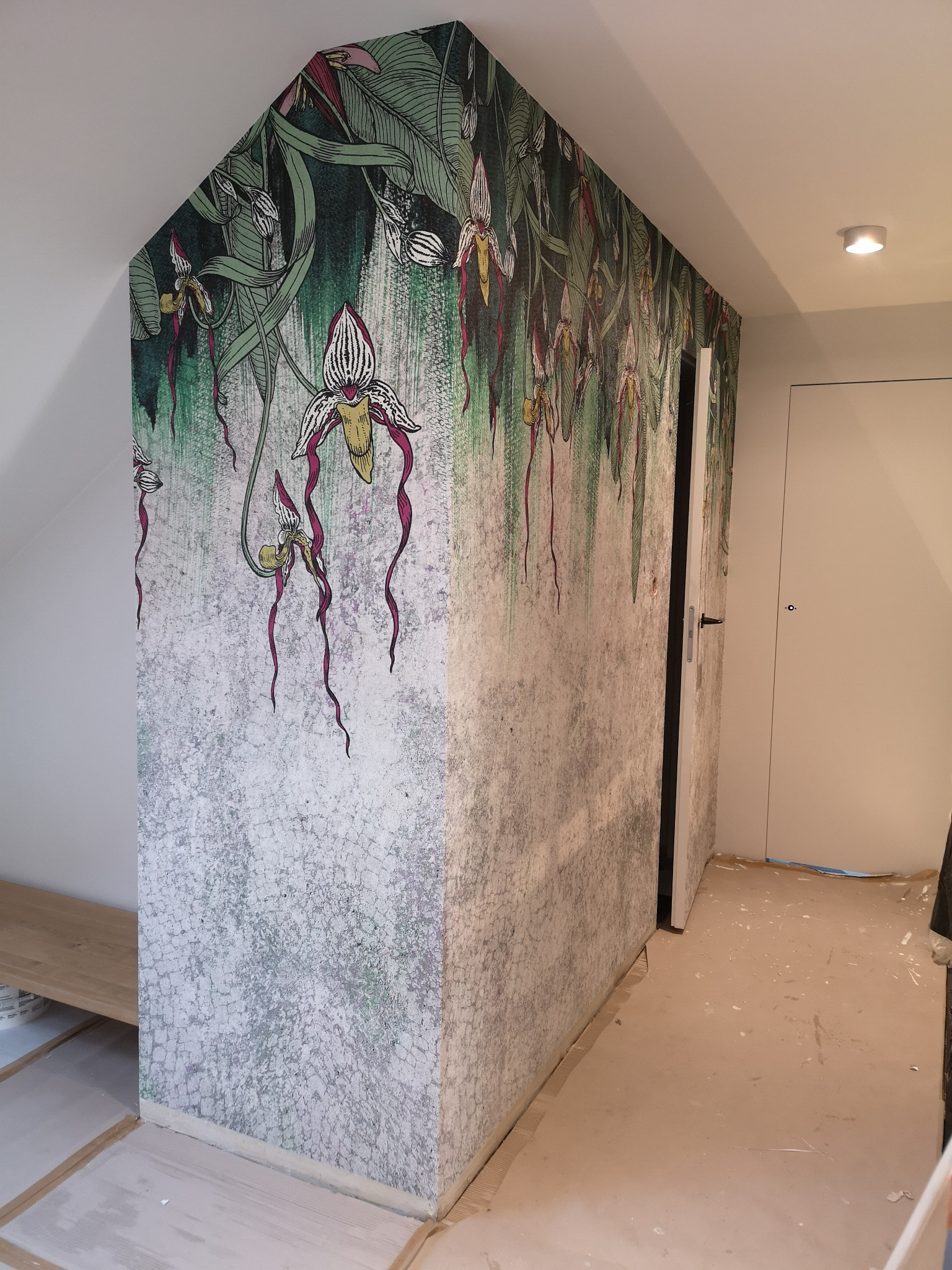Ściana w pokoju na poddaszu pokryta tapetą z motywem dżungli z egzotycznymi kwiatami; widoczne niedoskonałości wykończenia podłogi i listew przypodłogowych.