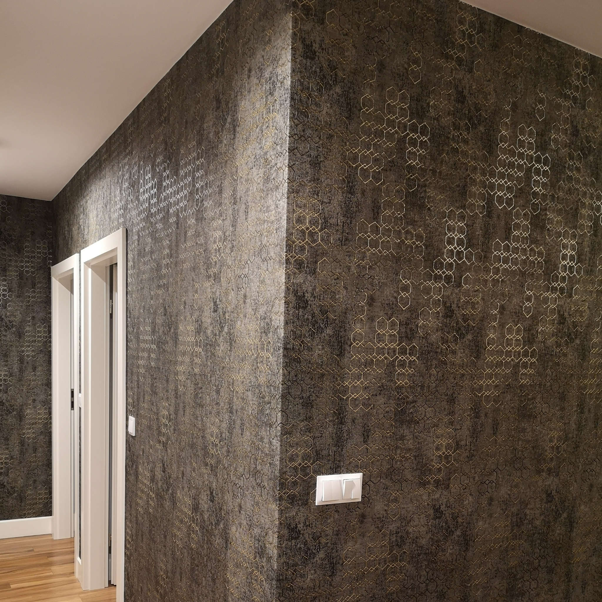Fragment ściany w korytarzu pokrytej tapetą w odcieniach brązu z geometrycznym wzorem w kształcie złotych sześciokątów, widoczne białe drzwi i włącznik światła.