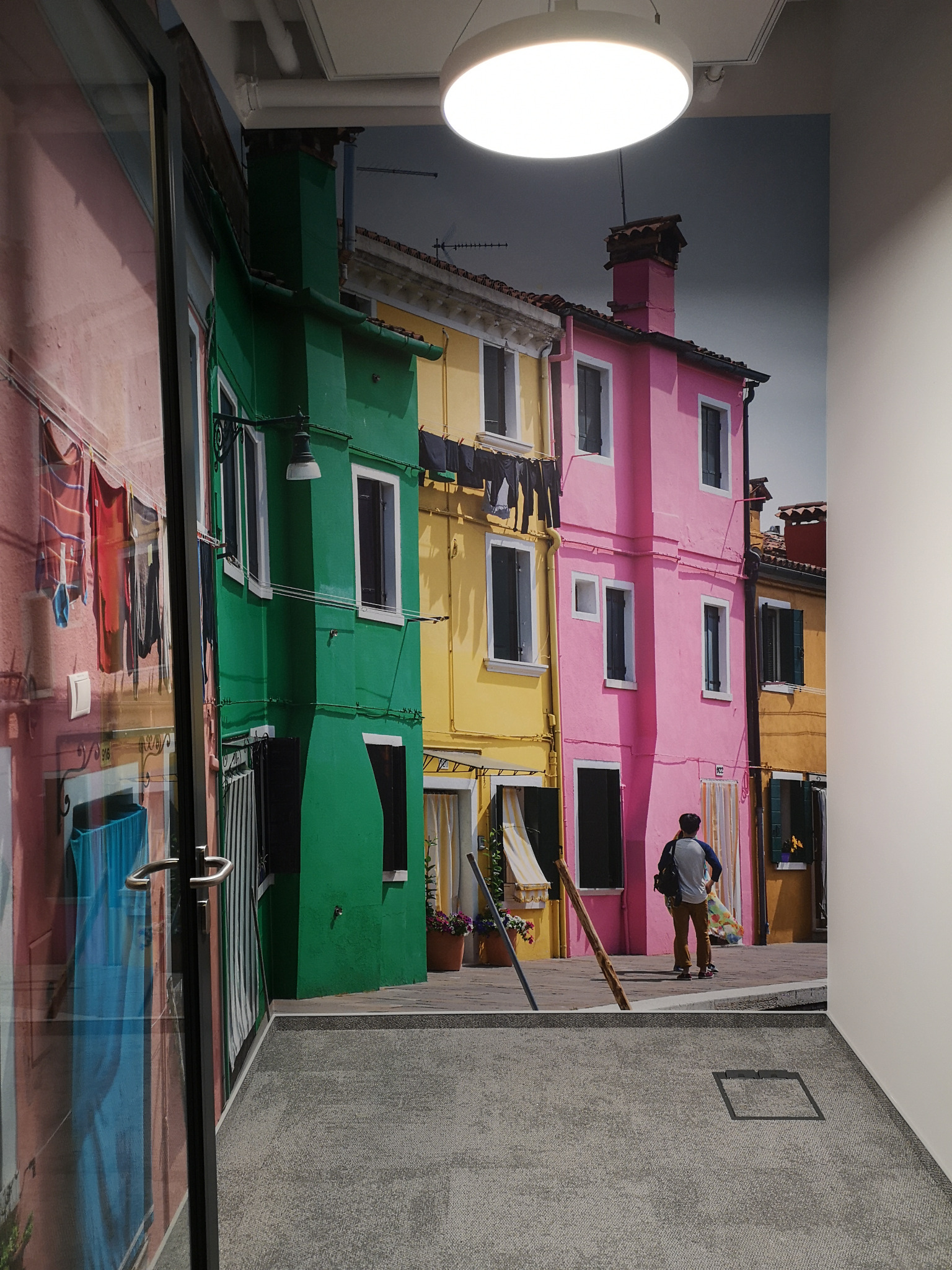 Fototapeta przedstawiająca kolorowe budynki w Burano, widoczna na ścianie biura z szarą wykładziną dywanową, odbicie w szklanej ścianie.