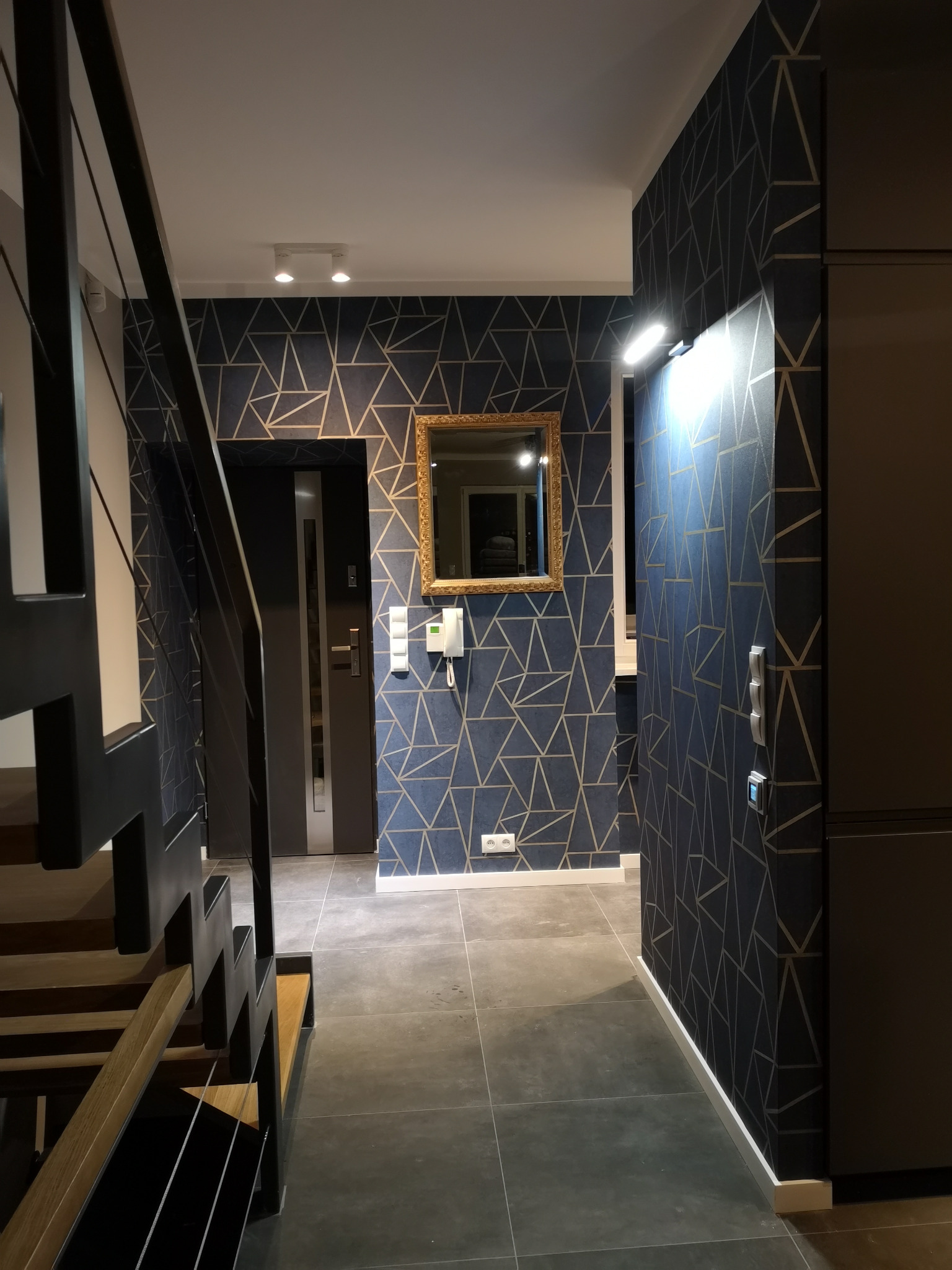 Wnętrze korytarza z granatową tapetą w geometryczne złote wzory, widoczne schody z drewnianymi stopniami i czarną metalową balustradą, lustro w złotej ramie na ścianie, szara podłoga z płytek.