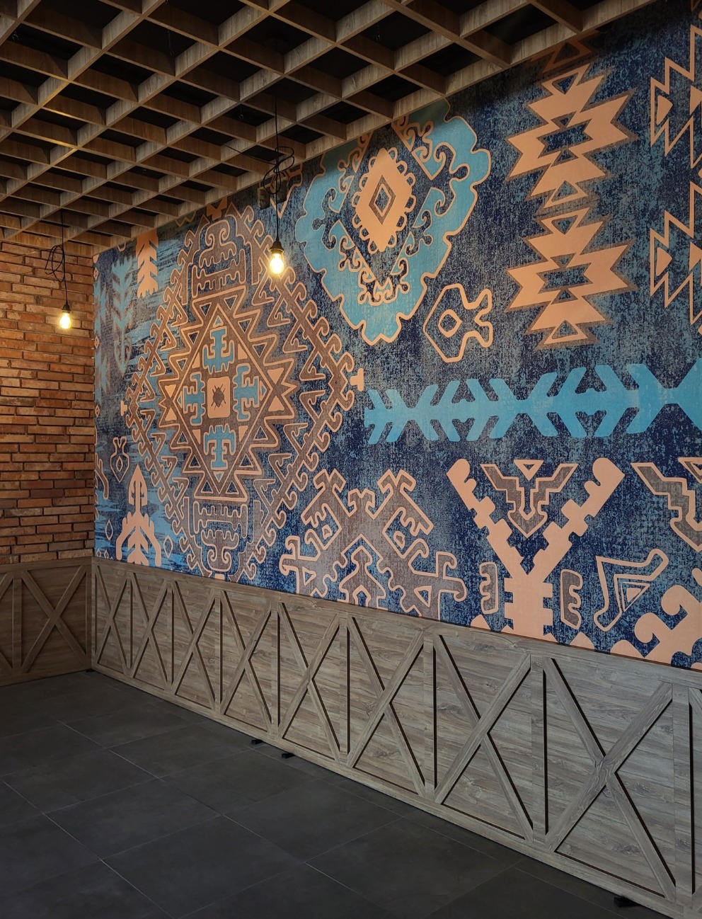 Wzorzysta tapeta w odcieniach błękitu i beżu z motywami etnicznymi, drewniana boazeria z geometrycznym wzorem rombów, ceglana ściana w rogu i sufit z drewnianych belek w restauracji.