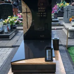 Nowoczesny, czarny pomnik z elementami beżowego kamienia i krzyżem, na cmentarzu; widoczna zapalona lampion i inne nagrobki w tle.