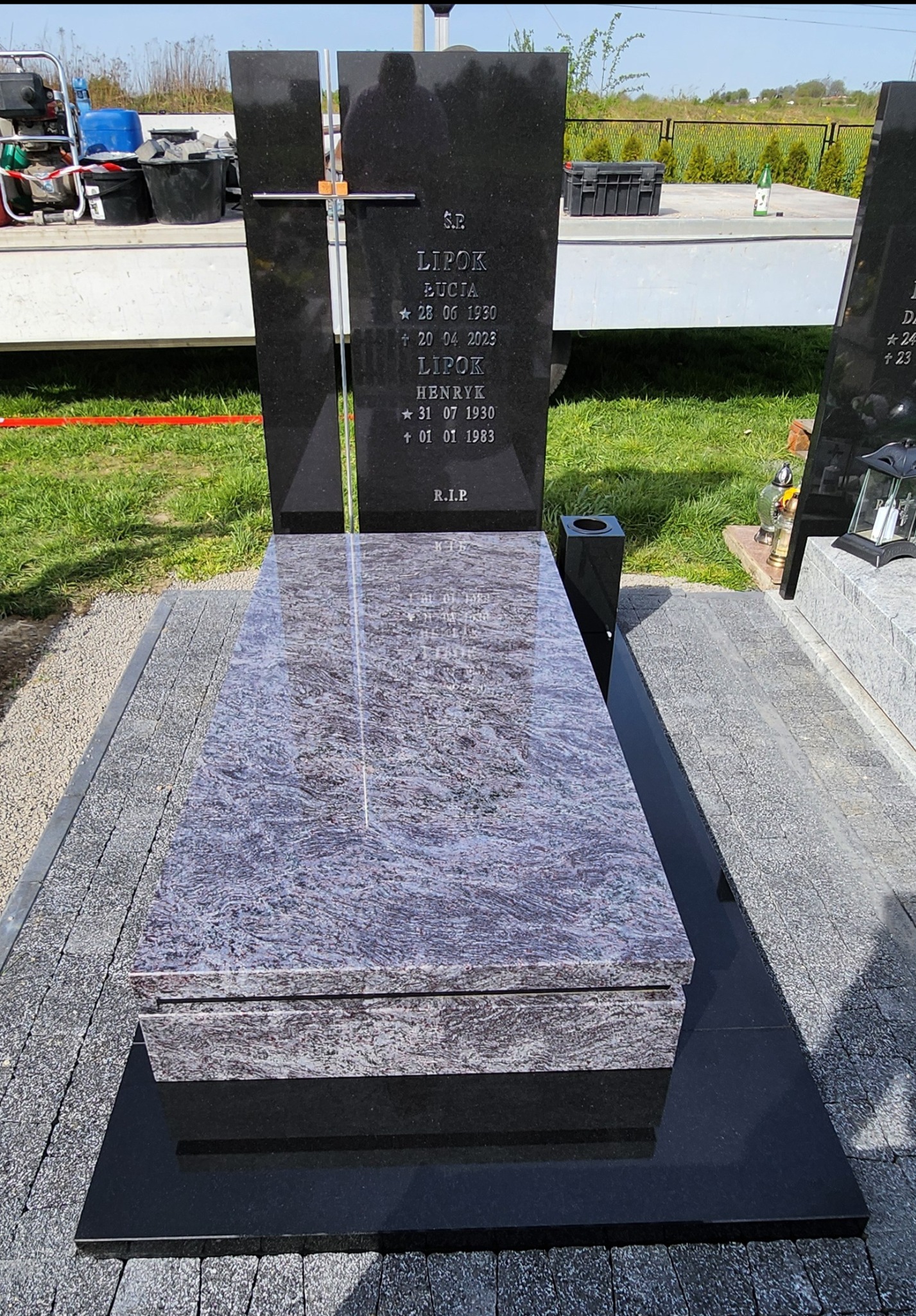 Czarny, podwójny pomnik z granitu z jasnoszarym nakryciem, otoczony szarą kostką brukową, na trawiastym cmentarzu.
