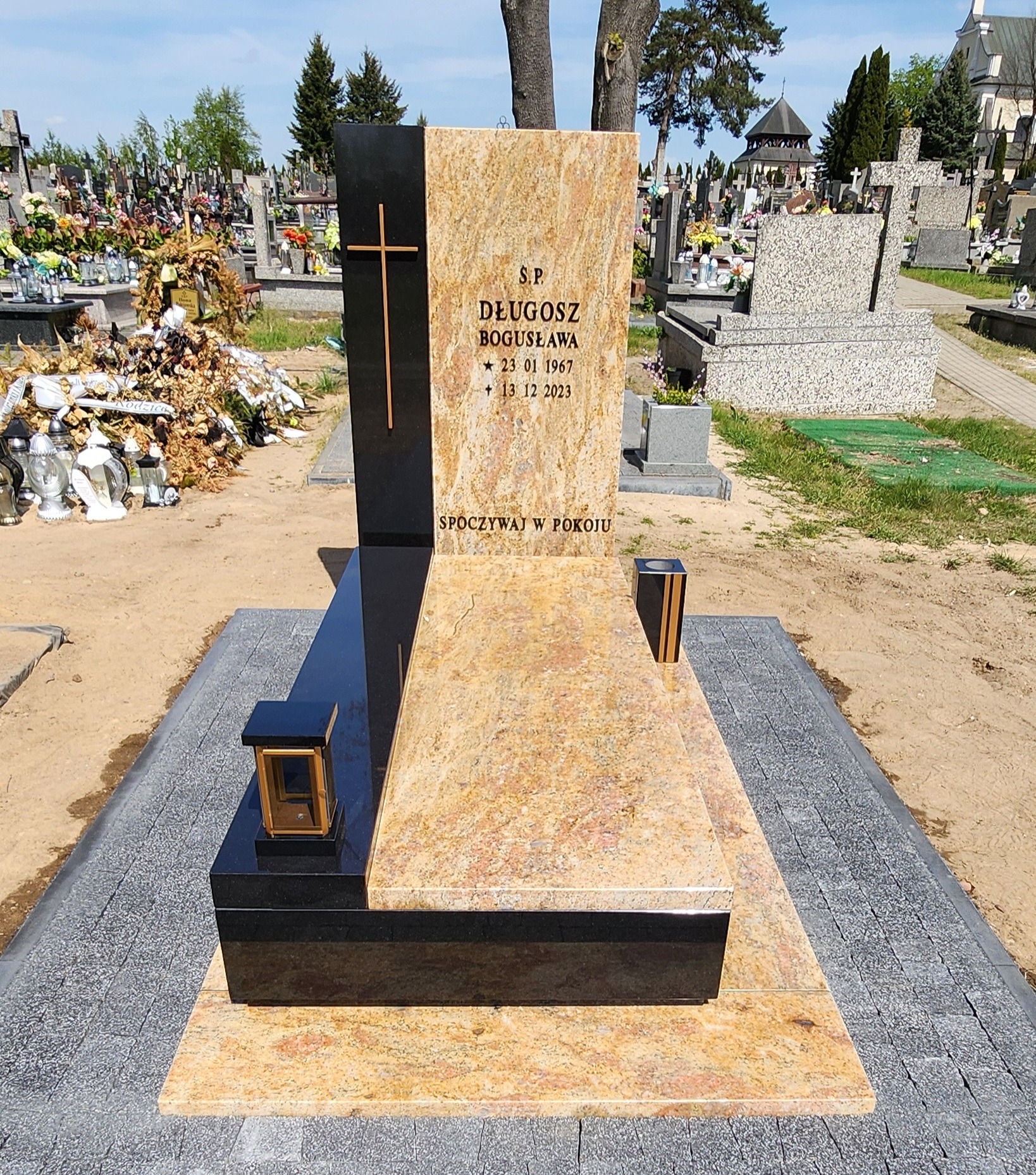Nowoczesny, dwukolorowy pomnik z jasnego i czarnego kamienia na cmentarzu, z wygrawerowanym krzyżem i danymi osoby zmarłej, otoczony szarą kostką brukową.