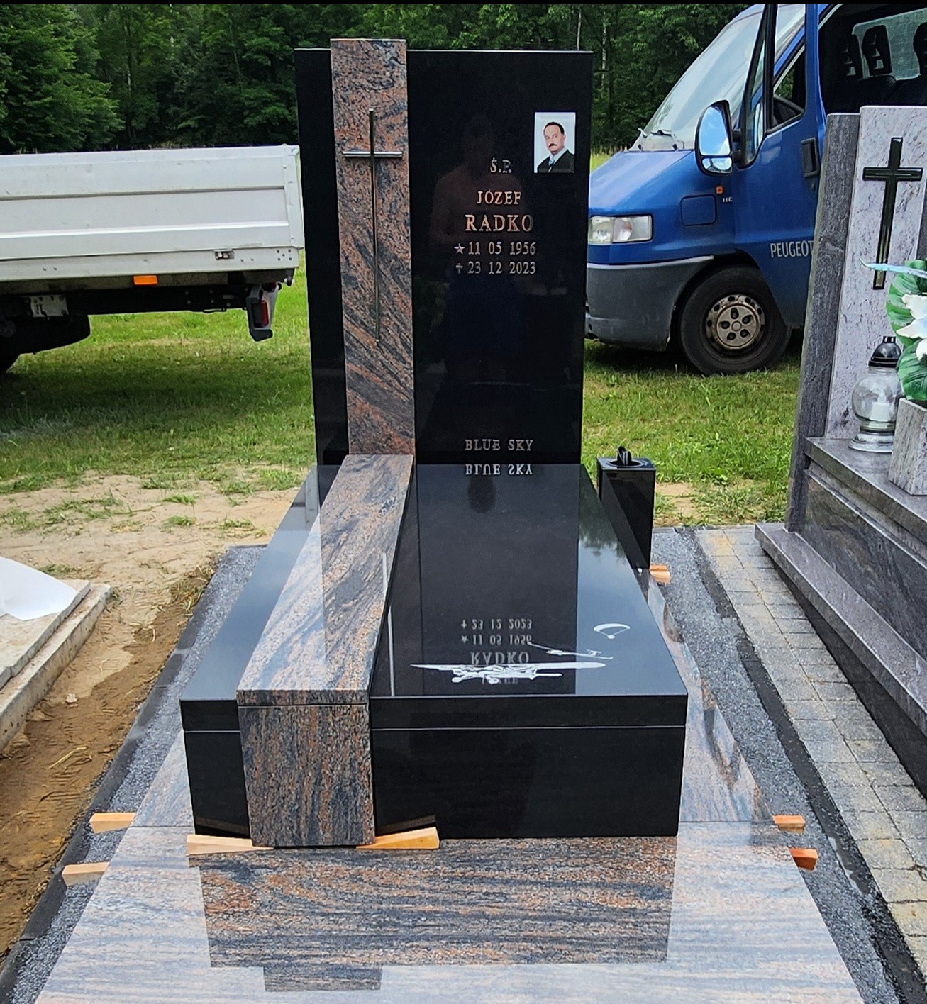 Czarny, nowoczesny pomnik z elementami z brązowego granitu, z wygrawerowanym wizerunkiem samolotu, na tle zaparkowanego niebieskiego vana.