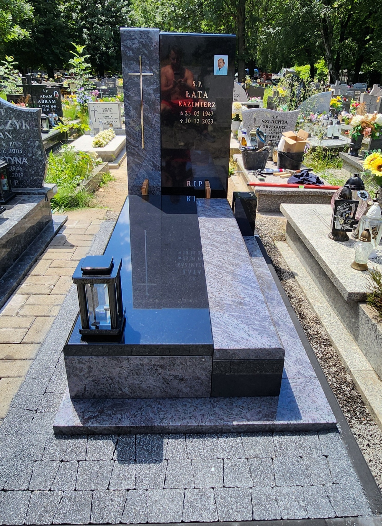 Nowoczesny, czarny nagrobek z elementami szarego granitu, z portretem zmarłego i datami urodzin oraz śmierci, ustawiony na cmentarzu z widocznymi innymi grobami w tle.