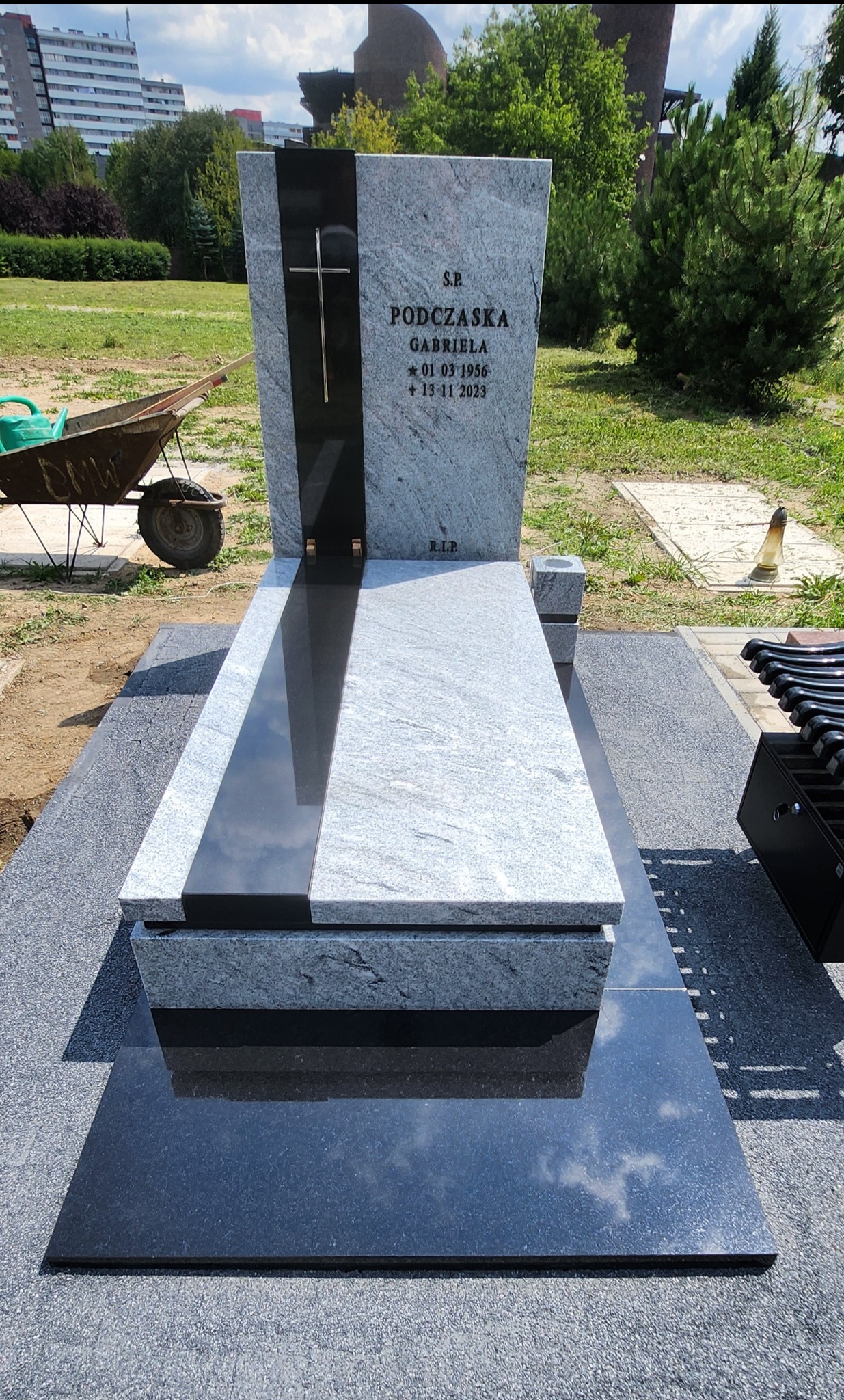 Jasnoszary pomnik z czarnym akcentem i wygrawerowanym krzyżem, otoczony zielenią parku i fragmentem wózka budowlanego z lewej strony.