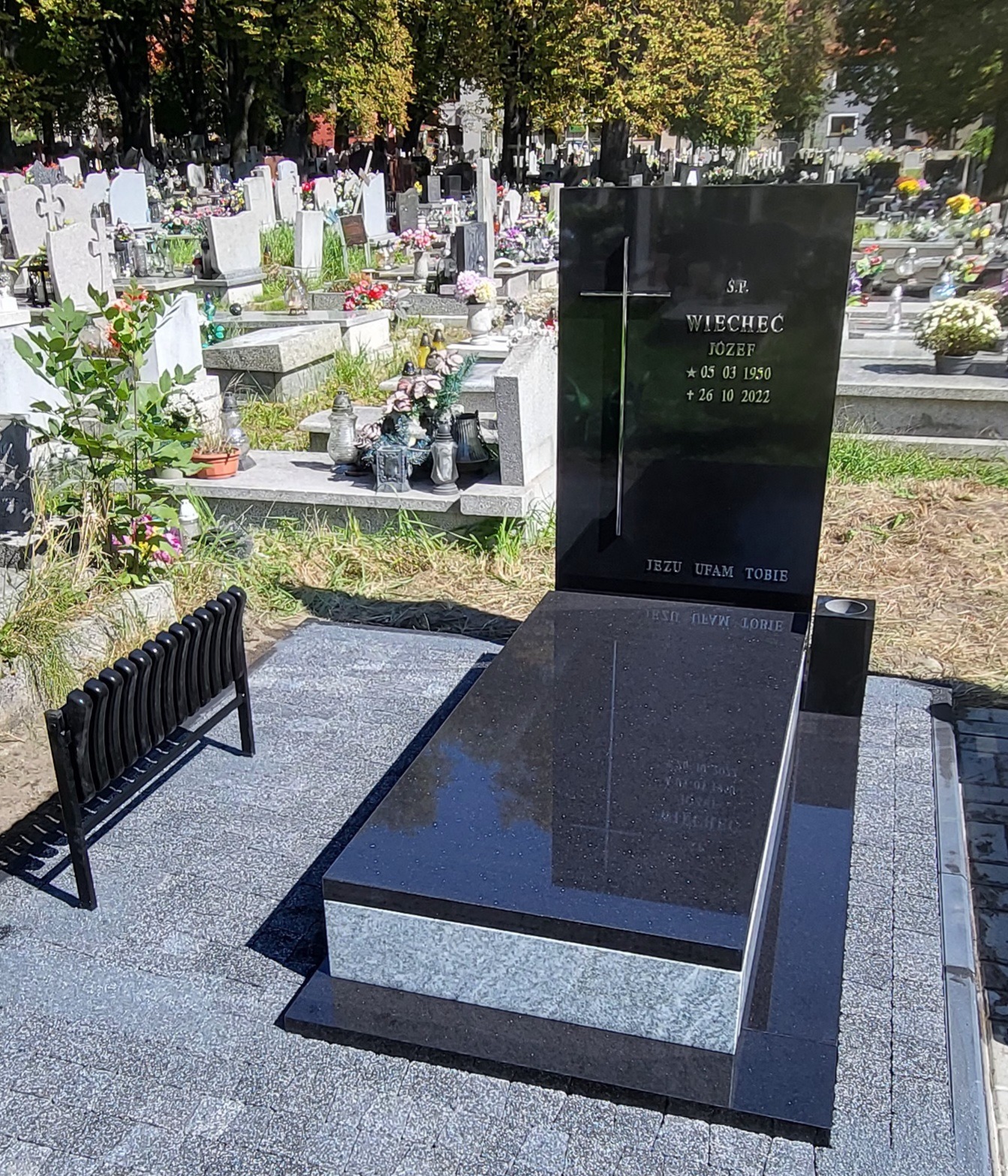 Czarny, polerowany pomnik z krzyżem i inskrypcją na cmentarzu, z małą czarną ławeczką obok. W tle inne groby i drzewa.